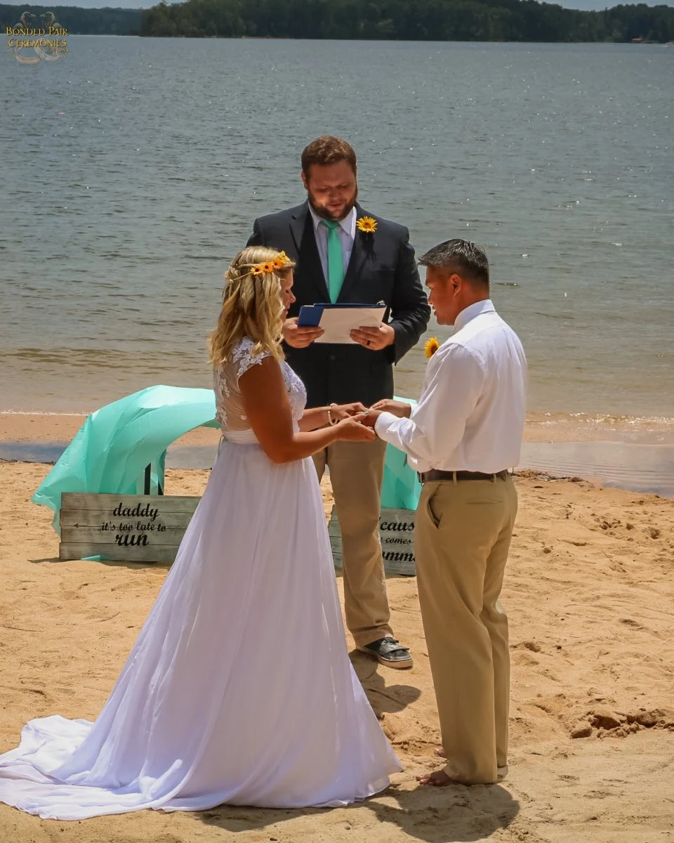 A couple getting married on a beach with an officiant overseeing the ceremony. The bride wears a white wedding dress and a flower crown, and the groom wears khaki pants and a white shirt. There are decorative signs and a teal umbrella in the background.