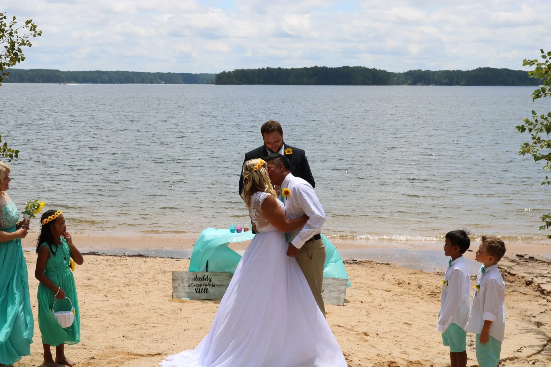 A wedding ceremony on a beach featuring a bride and groom sharing a kiss, with a wedding officiant and bridesmaids and groomsmen, all wearing coordinated attire in shades of aqua and white, by a lake with a scenic view of trees and water in the background.