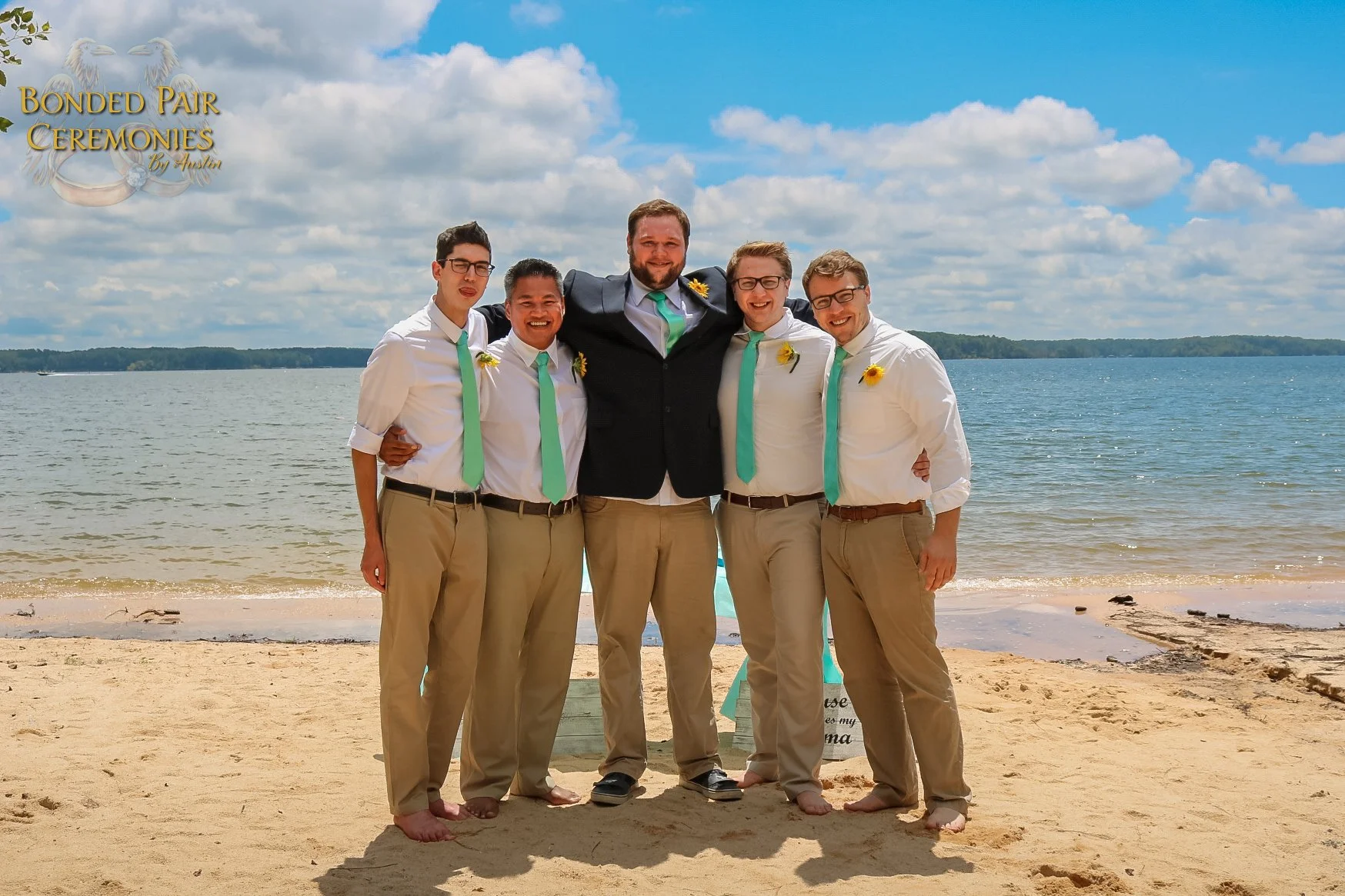 Group of six men standing on a sandy beach by the water, smiling and posing for a photo, with a cloudy blue sky in the background. They are dressed semi-formally with white shirts, beige pants, and turquoise ties, except for one man in a black suit jacket. Some wear glasses and have small yellow flowers pinned to their shirts.