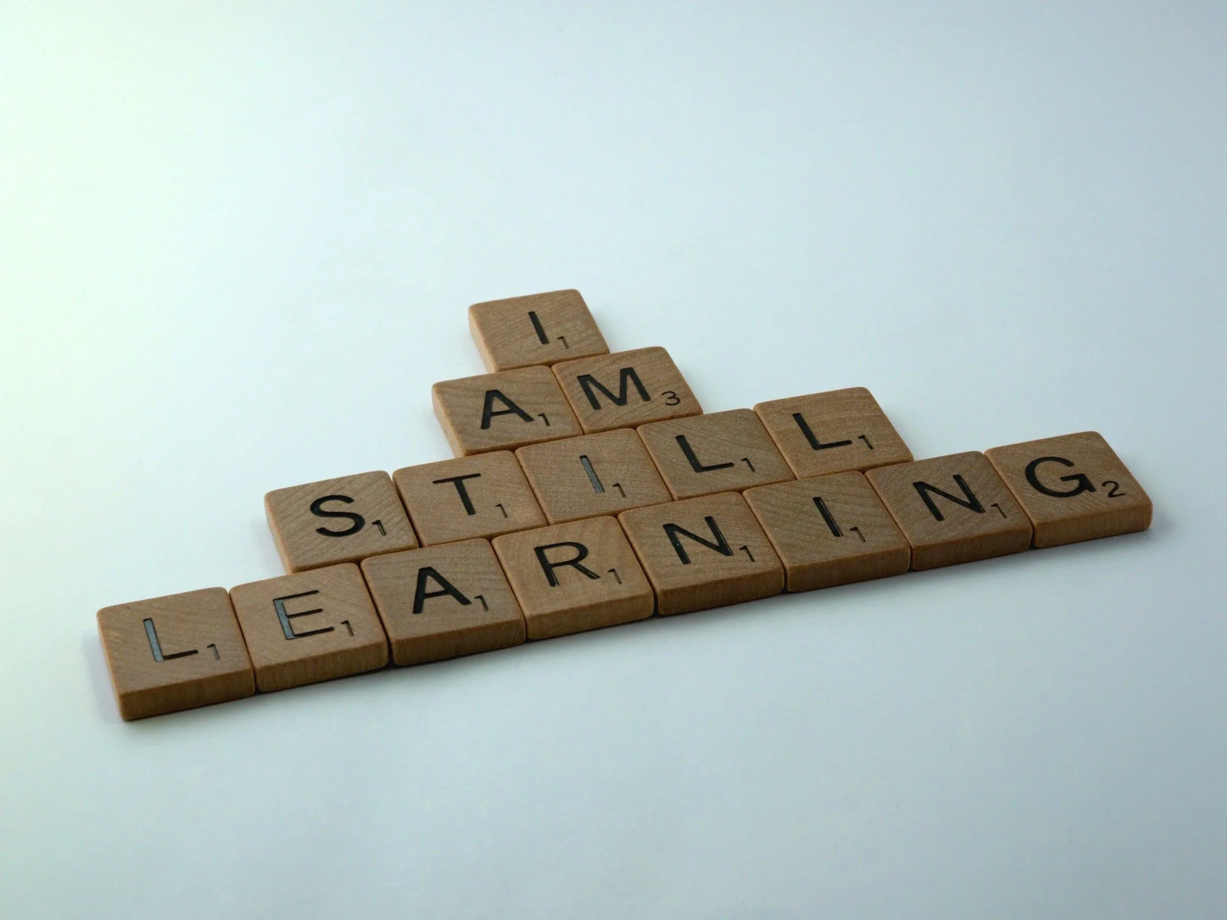 Wooden Scrabble tiles arranged to spell the phrase 'I AM STILL LEARNING' on a white background symbolising the process of learning with guidance.
