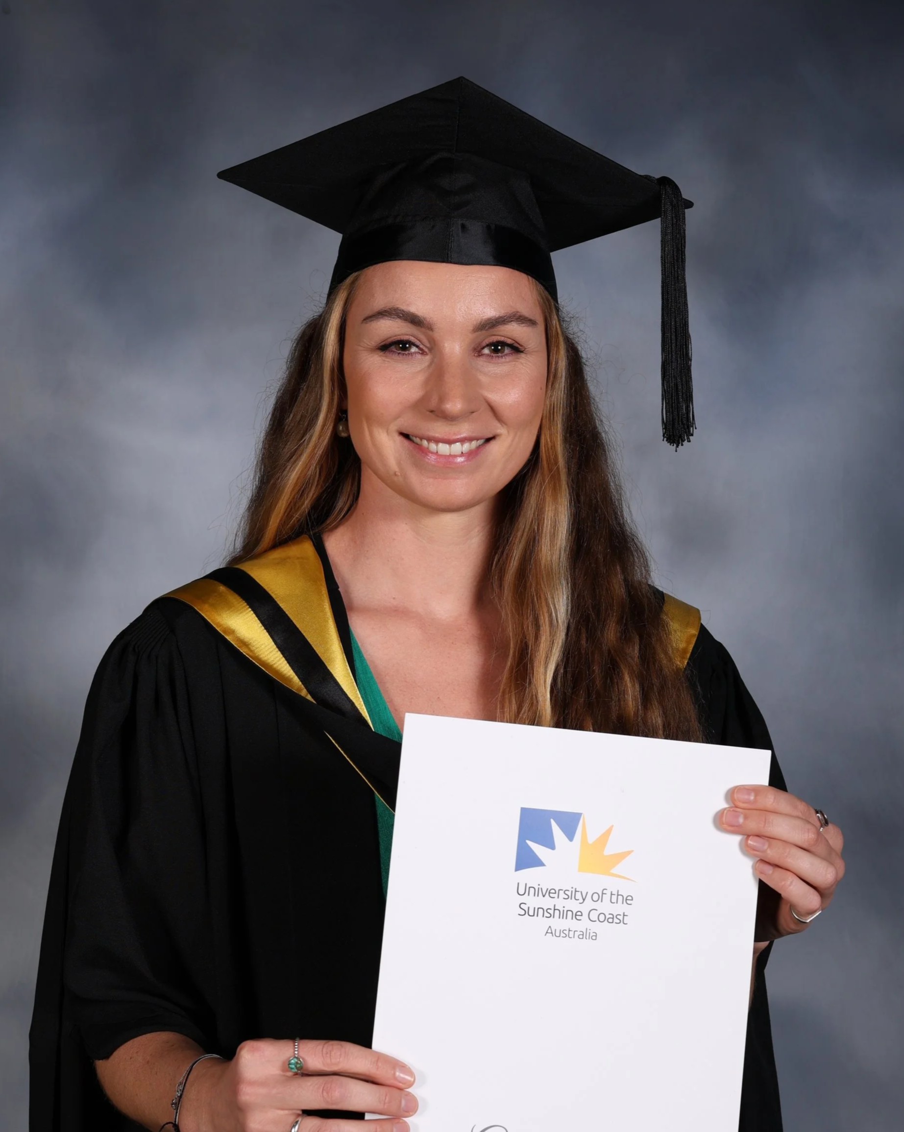 A woman in graduation attire holding a diploma from the University of the Sunshine Coast, Australia, smiling at the camera.