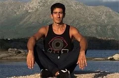 Hombre sentado en la orilla del agua con montañas en el fondo, vistiendo ropa deportiva negra.