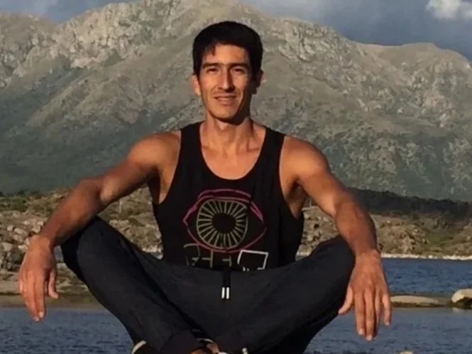 Hombre sentado en una roca cerca de un cuerpo de agua con montañas al fondo.