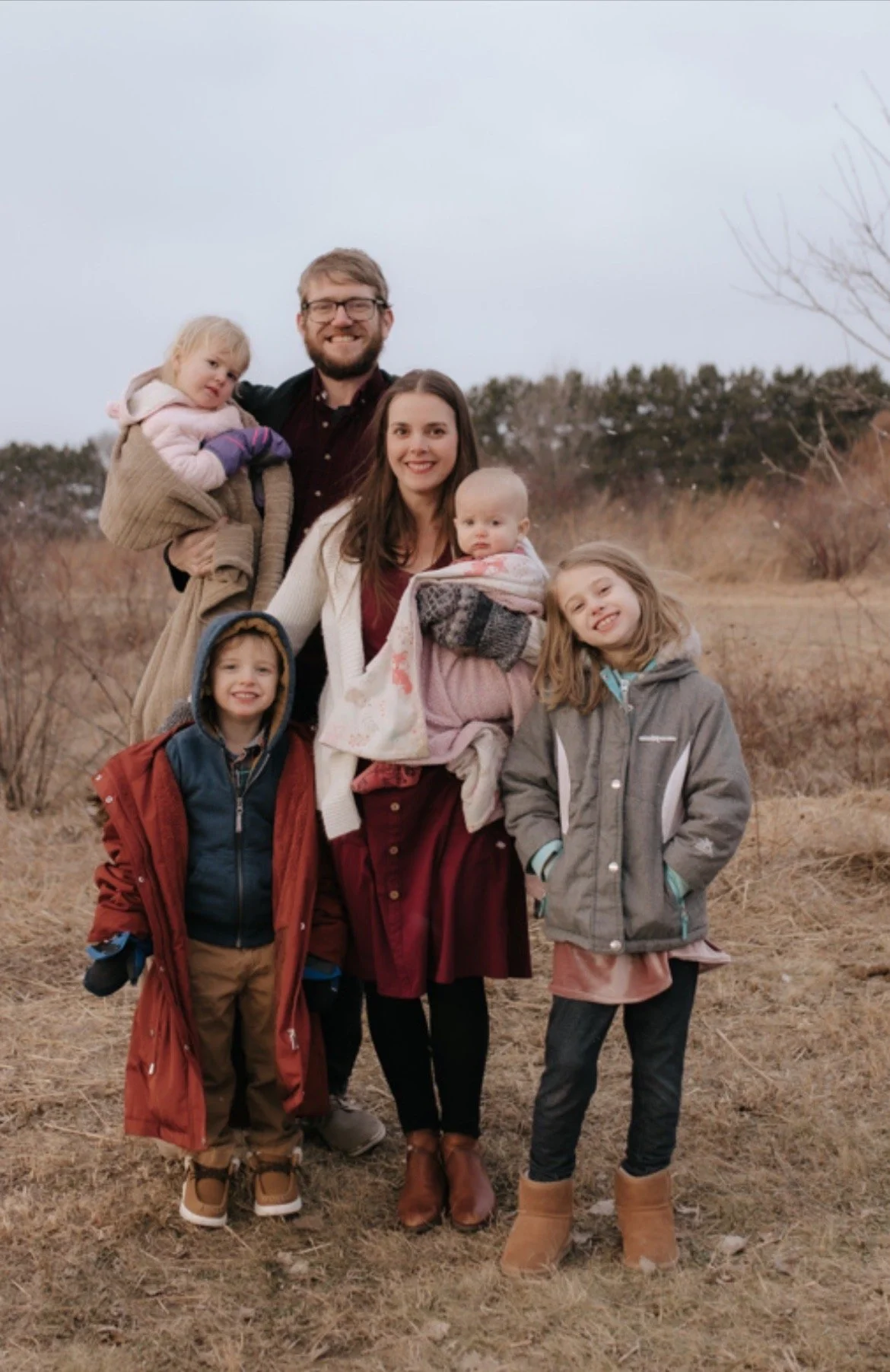 Family of six outdoors during fall or winter, standing in a field with dry grass and bushes, wearing warm clothing.