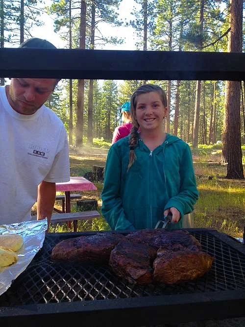 Girl smiling near a charcoal grill with cooked steaks, in a wooded outdoor area.