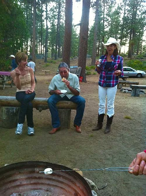 Three people outdoors in a wooded area, two women and one man, are socializing around a campfire. One woman in a plaid shirt and white cowboy hat is holding drinks, while the man in the center is eating. A woman on the left sits on a log, and there a