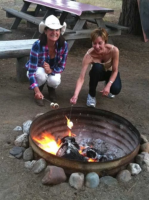 Two women roasting marshmallows over an outdoor campfire at a picnic area, with picnic tables in the background.