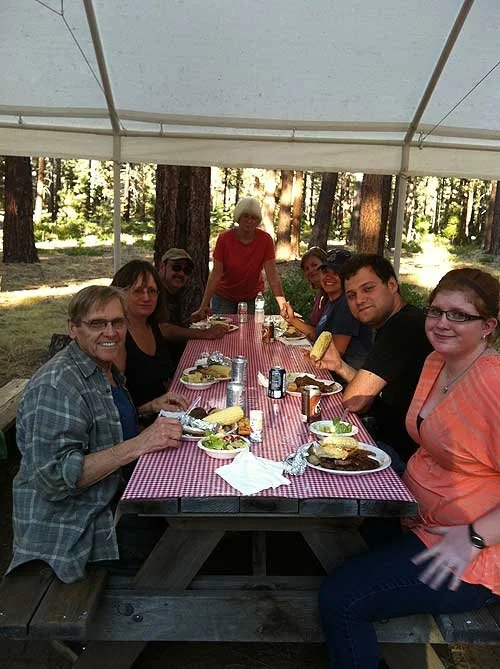 Group of seven people sitting at a picnic table outdoors under a white canopy, enjoying a meal with trees in the background.