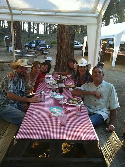 Group of six people sitting around a picnic table under a canopy outdoors, with trees and parked cars in the background, enjoying a meal with drinks and food on a red checkered tablecloth.