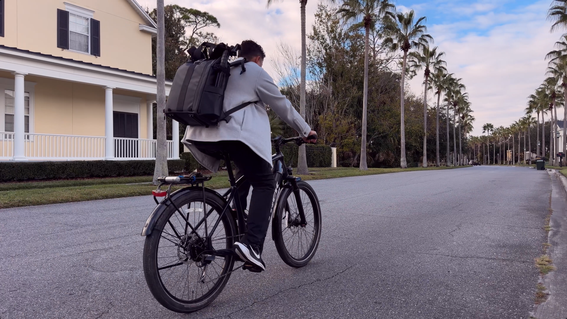 A person riding a black bicycle on a residential street with tall palm trees lining the sidewalk and a yellow house with white pillars in the background.