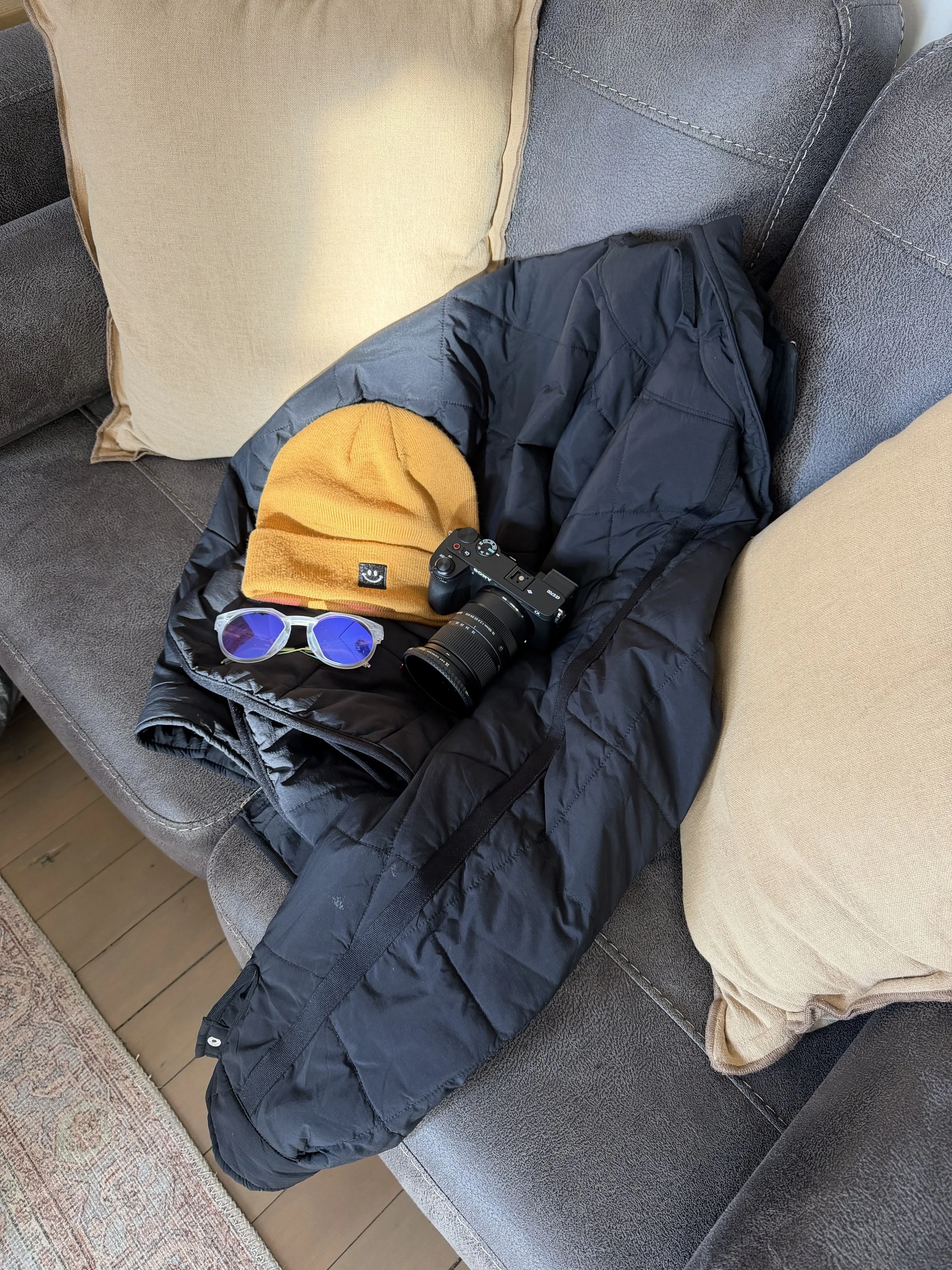 A black jacket, yellow beanie, blue sunglasses, and a camera are arranged on a black sleeping bag on a gray couch, with beige pillows behind.