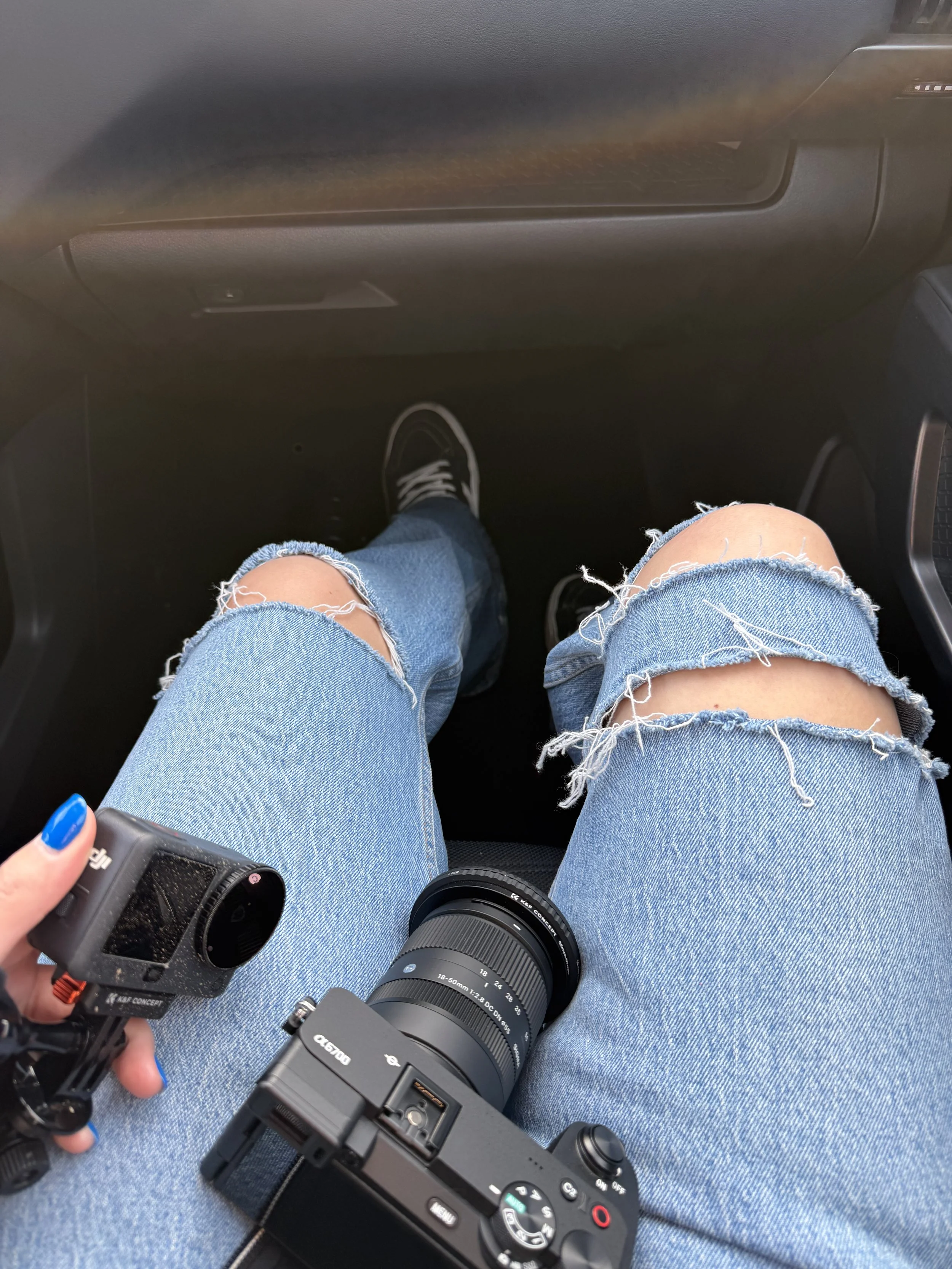 A person wearing ripped blue jeans is sitting in a vehicle, holding a black camera with a zoom lens, with part of their fingernail painted blue visible in the corner.