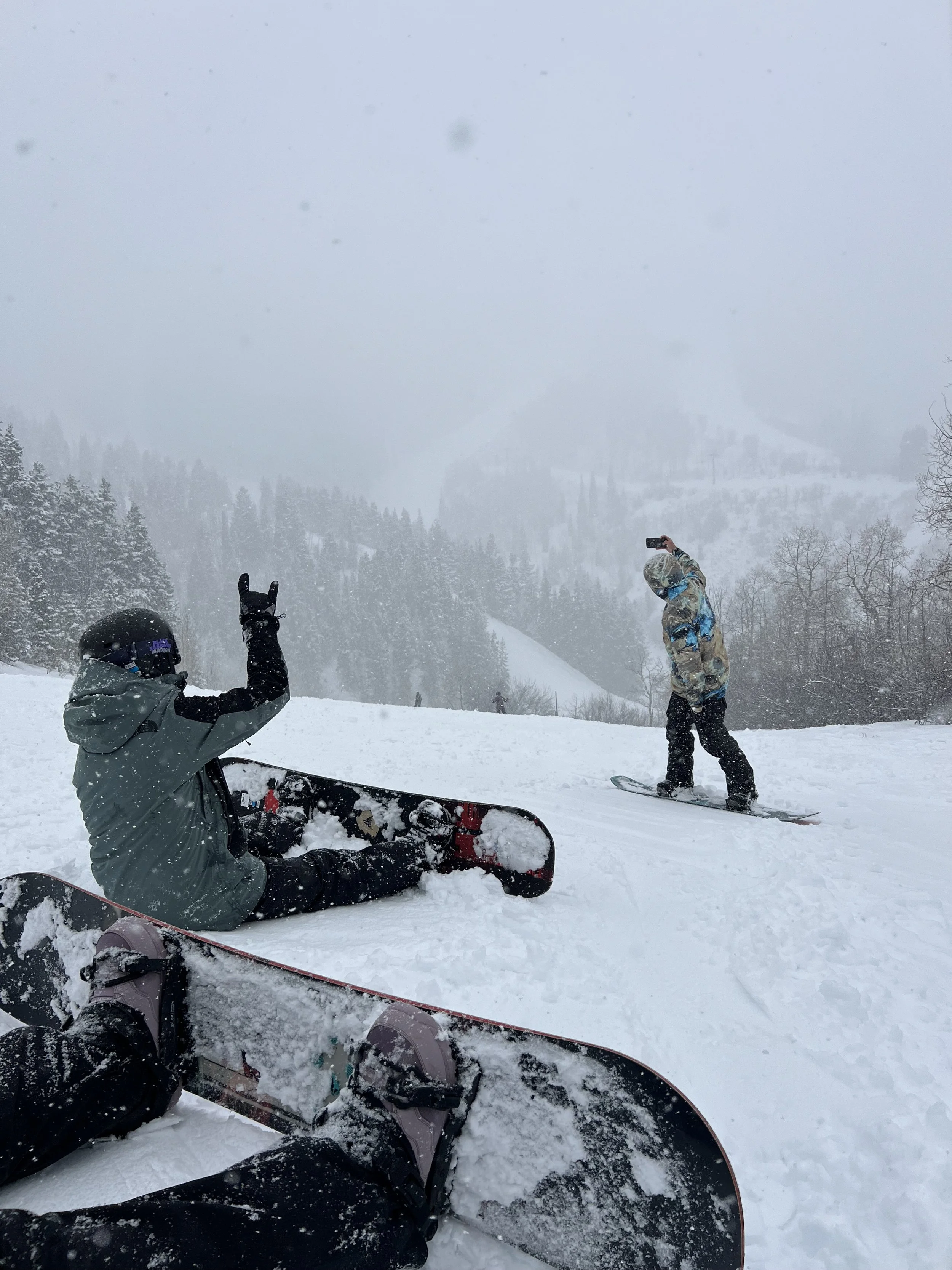 People in winter gear snowboarding and taking photos on a snowy mountain with trees in the background.
