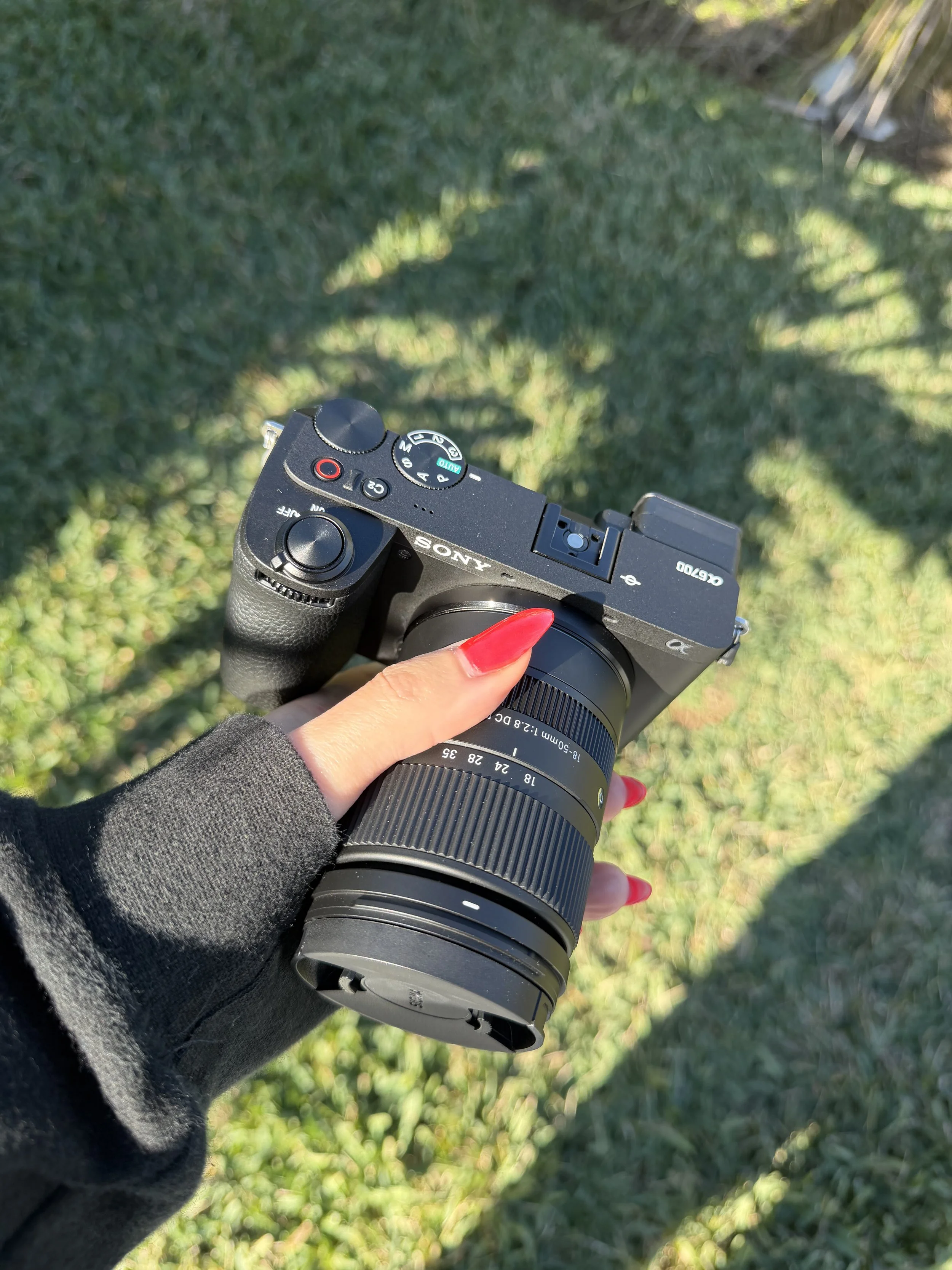 A person holding a Sony camera with a telephoto lens, wearing a black long sleeve top, standing outdoors on grass.