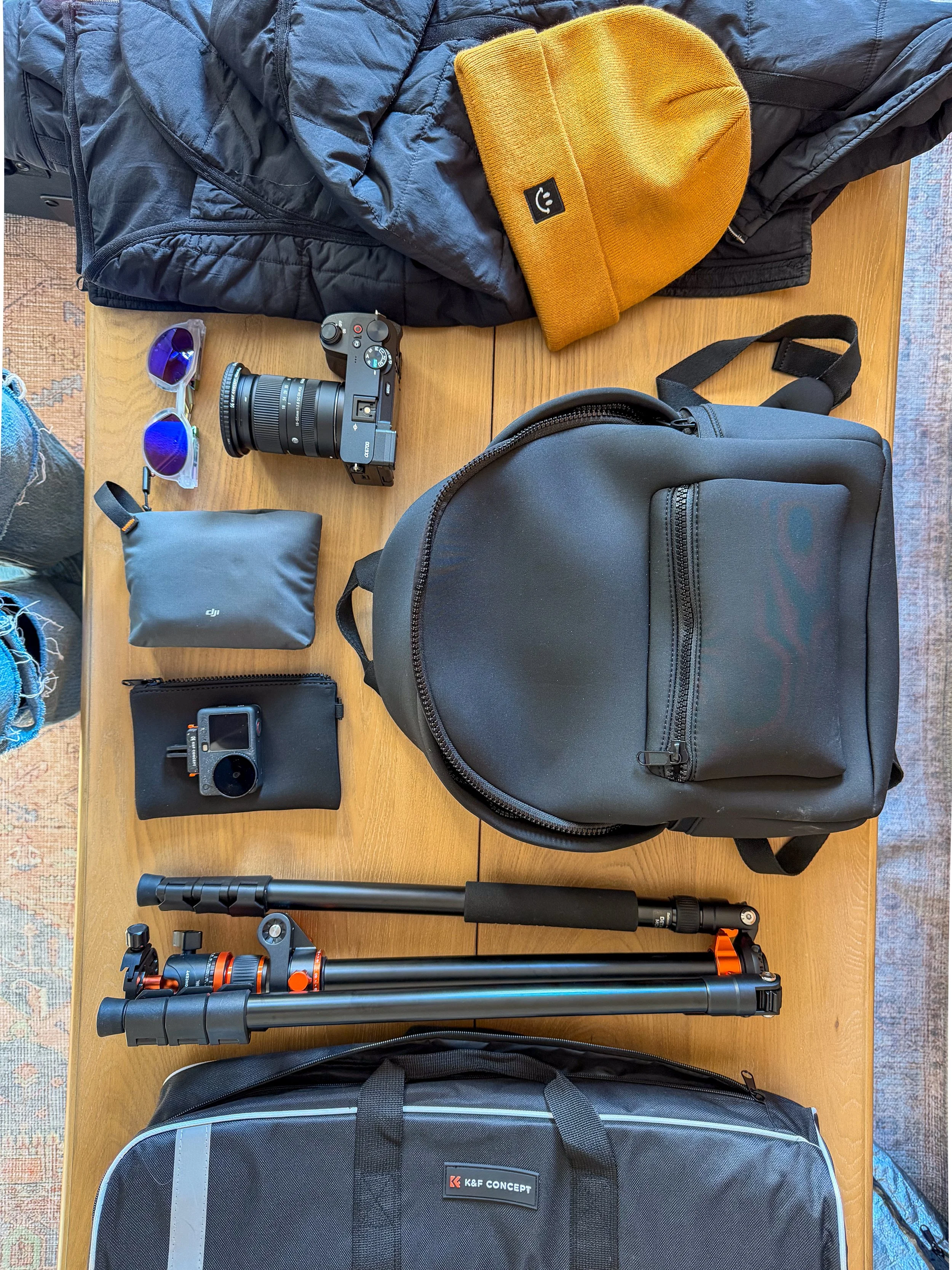 Photography gear on a wooden table, including a black backpack, a camera with a lens, purple sunglasses, a yellow beanie, a black padded jacket, a small black drone with controller, trekking pole, tripod, and a gray K&F Concept bag.