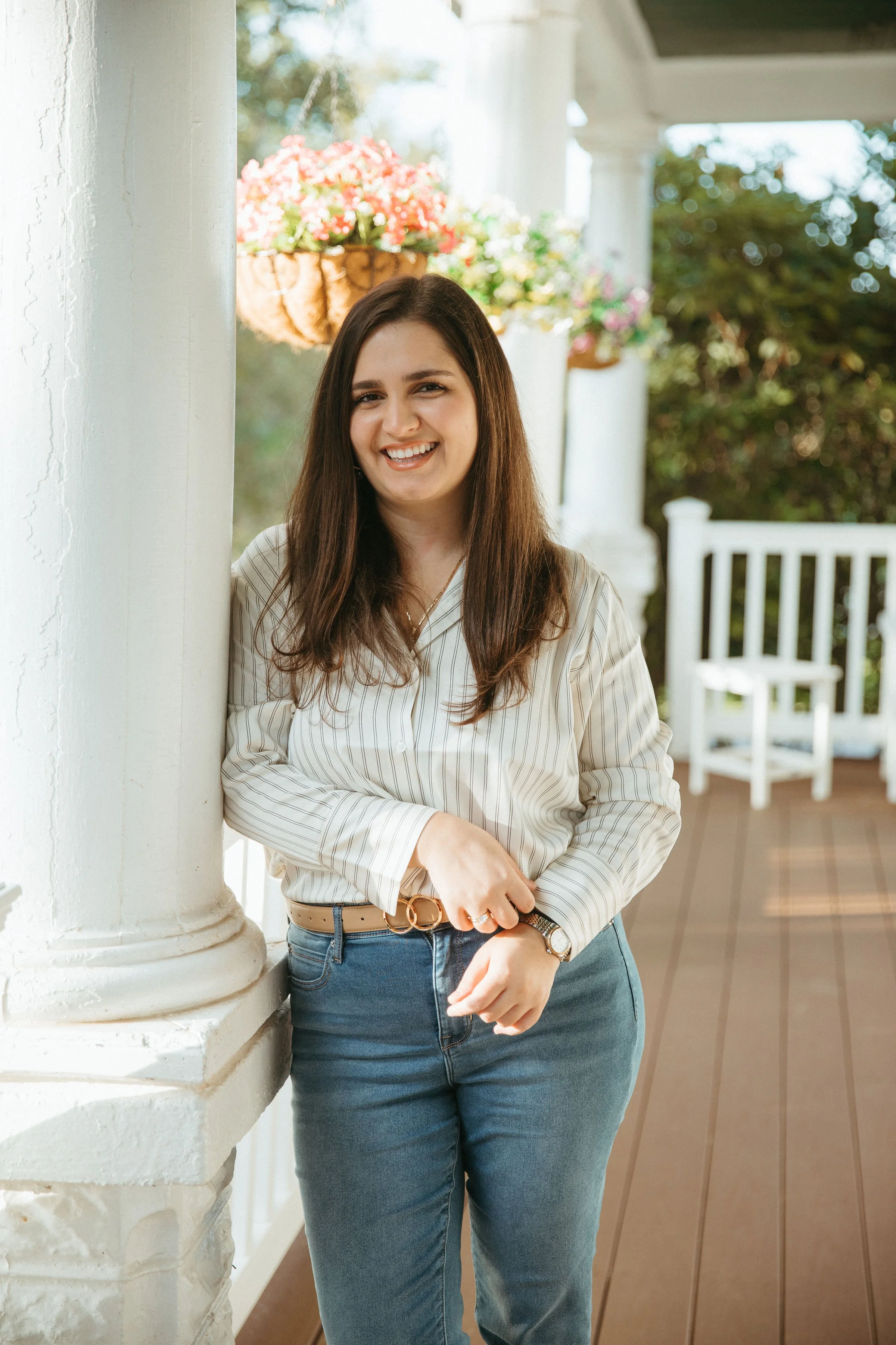 A young woman with long brown hair, smiling, standing on a porch with white columns and hanging flower baskets, wearing a striped button-up shirt and blue jeans.