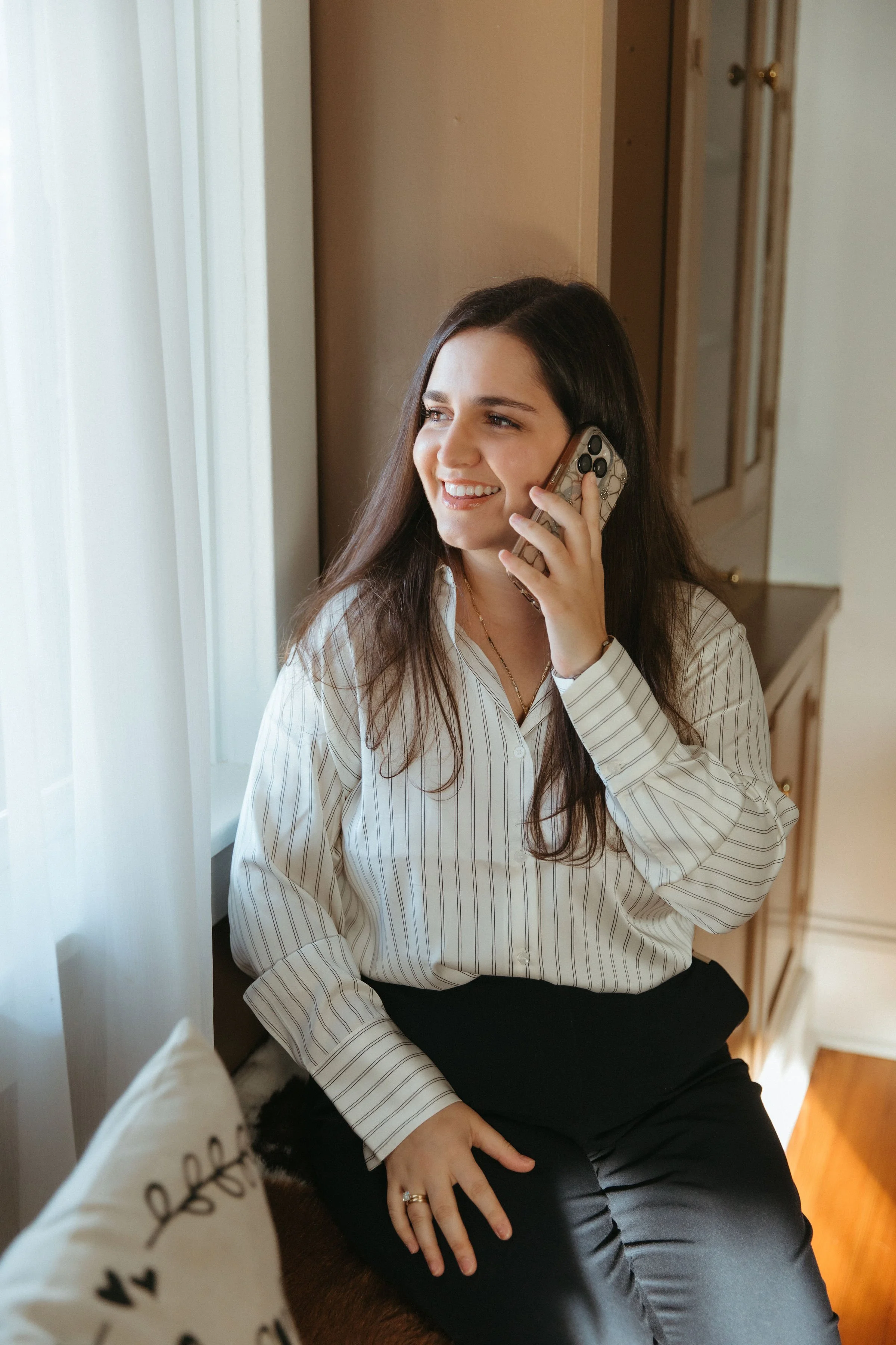 A woman with long brown hair is sitting by a window, smiling and talking on a smartphone.