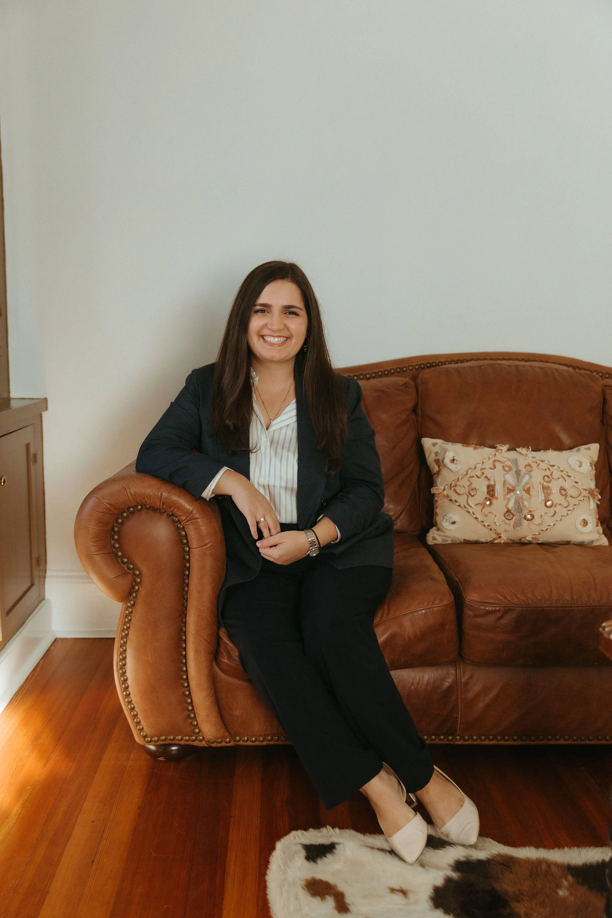 A woman with long dark hair, dressed in a dark blazer, white blouse, and black pants, sitting on a brown leather couch smiling at the camera.