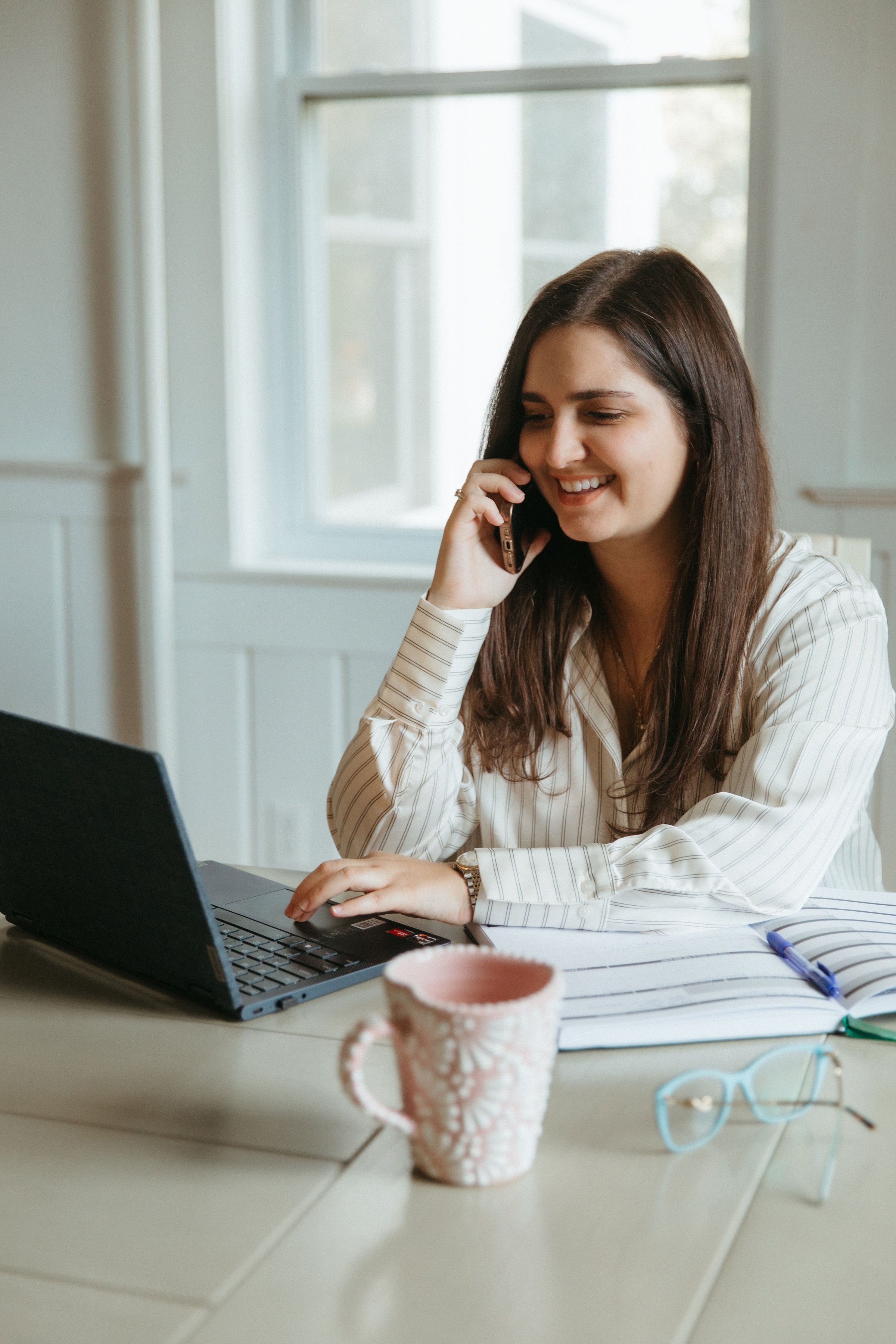 A woman with long brown hair smiling while talking on her mobile phone at a desk with a laptop, papers, a blue pen, pink and white coffee mug, and glasses, in a bright room with large windows.