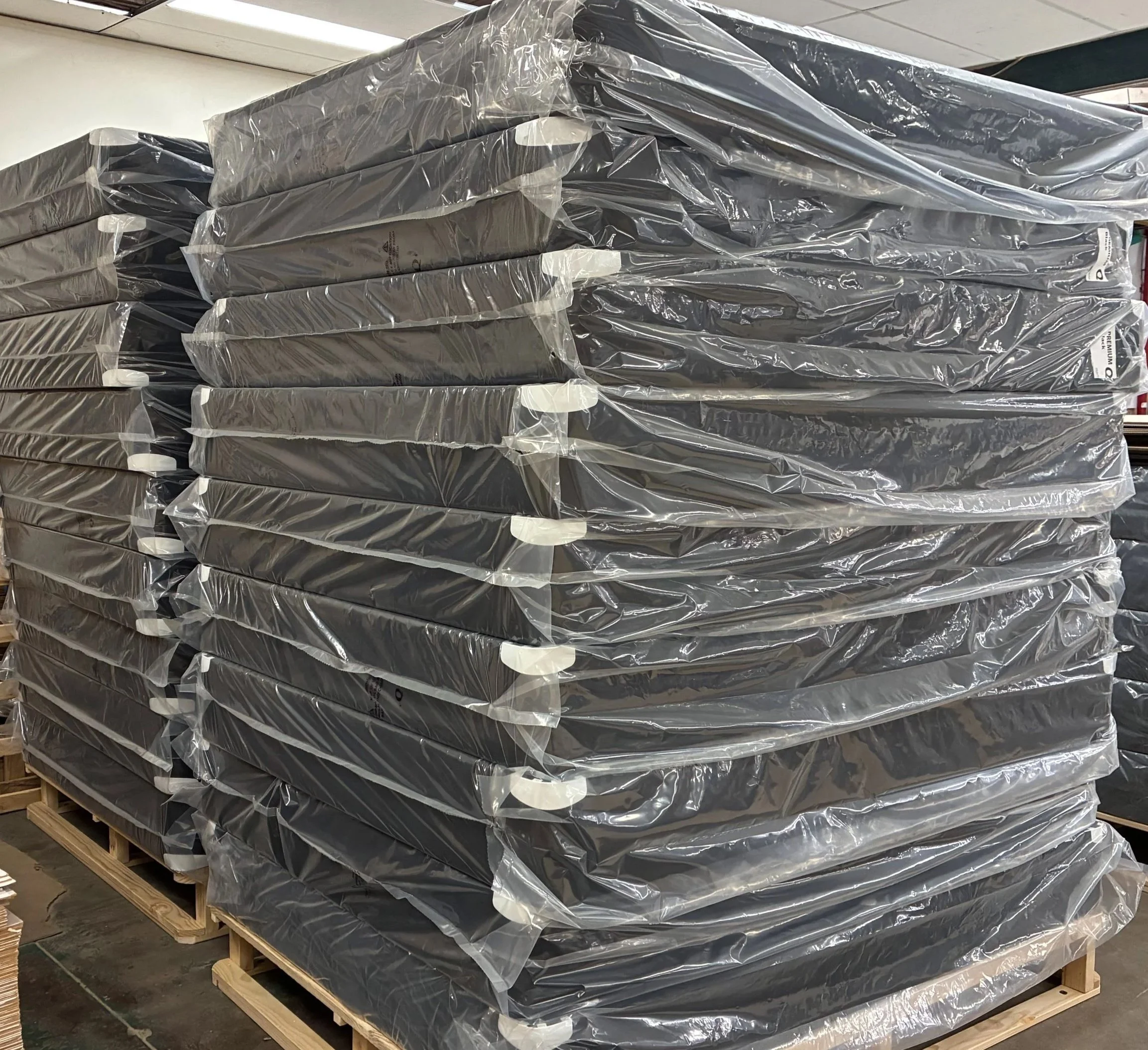 Stacks of packaged black bed bases wrapped in plastic on wooden pallets in a warehouse.