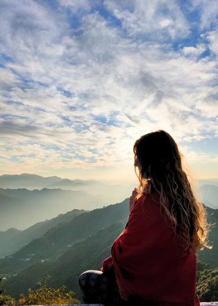 Woman sitting on a mountain with her back to the camera, overlooking a scenic view of rolling green mountains and a partly cloudy sky at sunrise or sunset.