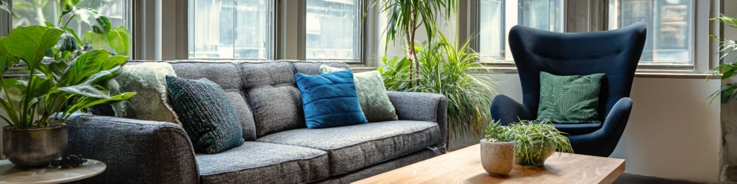 An office space with a gray sofa, blue and textured throw pillows, a blue armchair with a green pillow, and several potted plants near large windows.