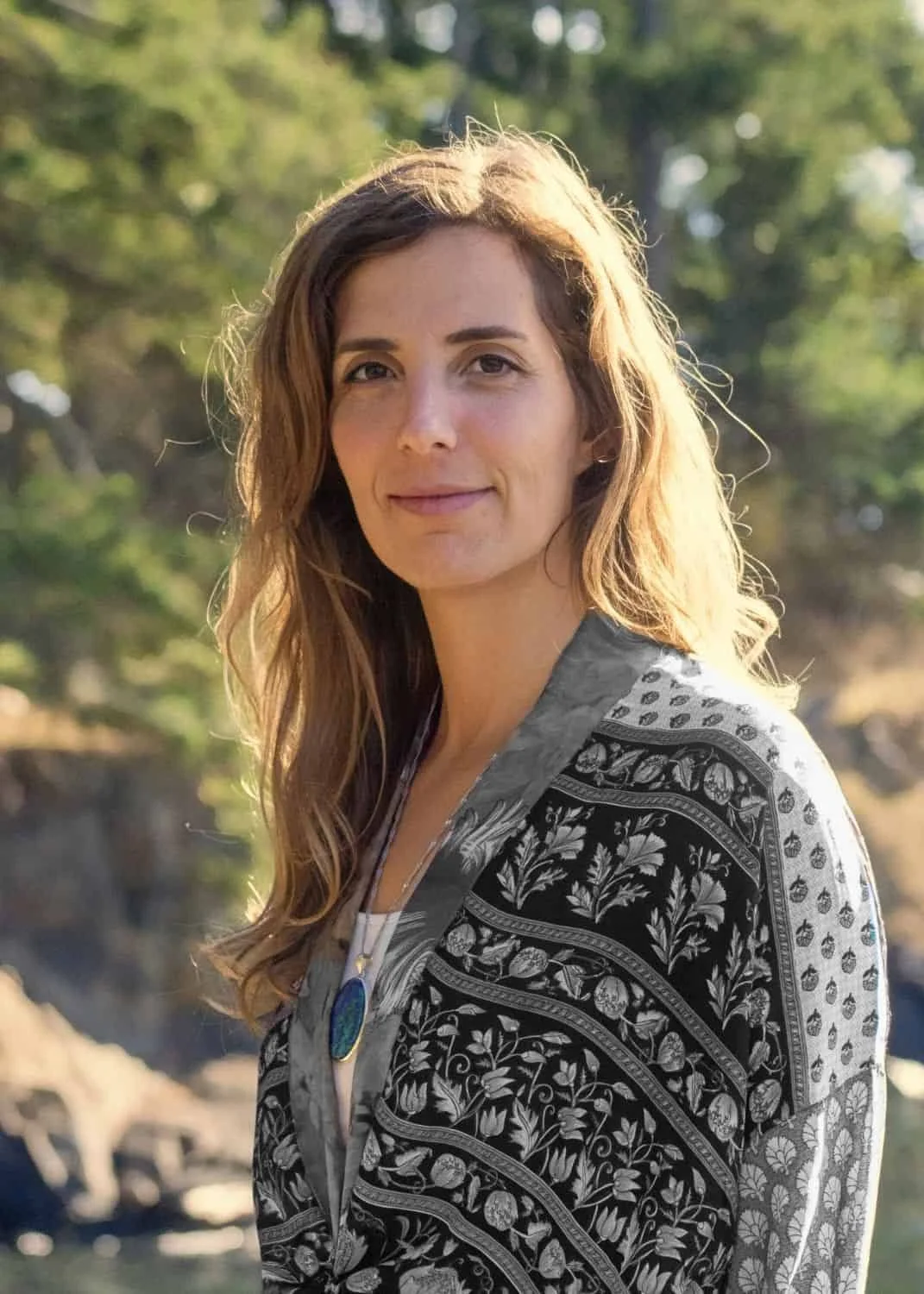 A woman with long, wavy brown hair and light skin standing outdoors in front of trees, smiling slightly, wearing a gray patterned jacket and a necklace with a large oval blue pendant.