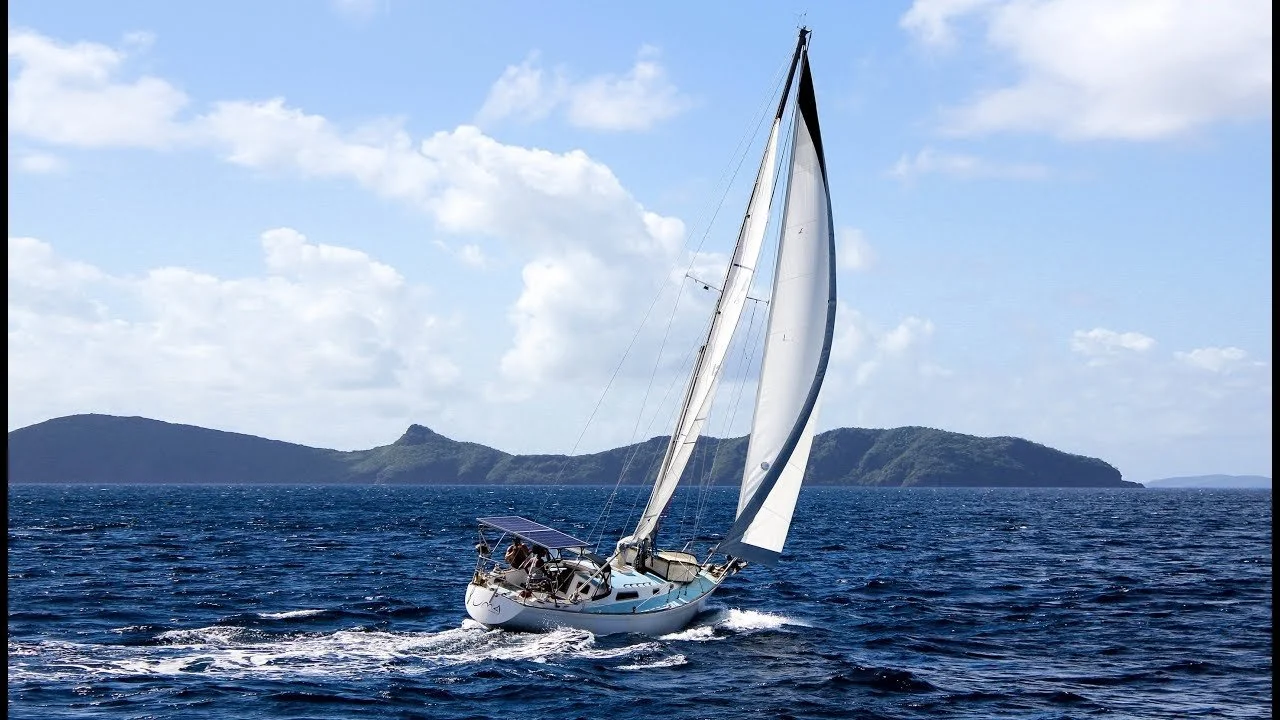 A sailboat with white sails on the ocean with a distant island and blue sky with clouds. The image underscores the advantages of a charted investment plan via the bucket investing principles outlined on the site.