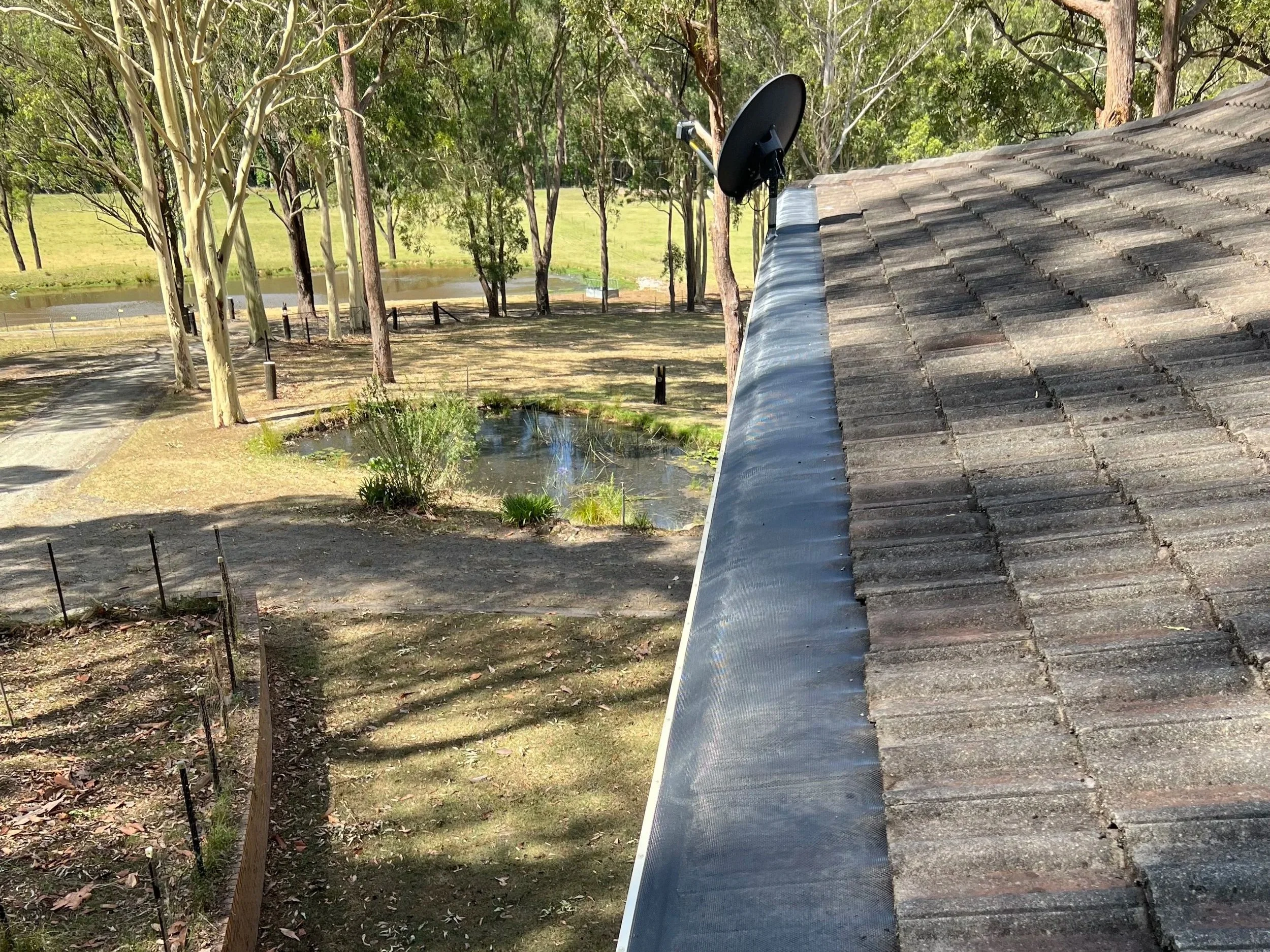 A rooftop with gray shingles and a clear gutter overlooking a park with trees.