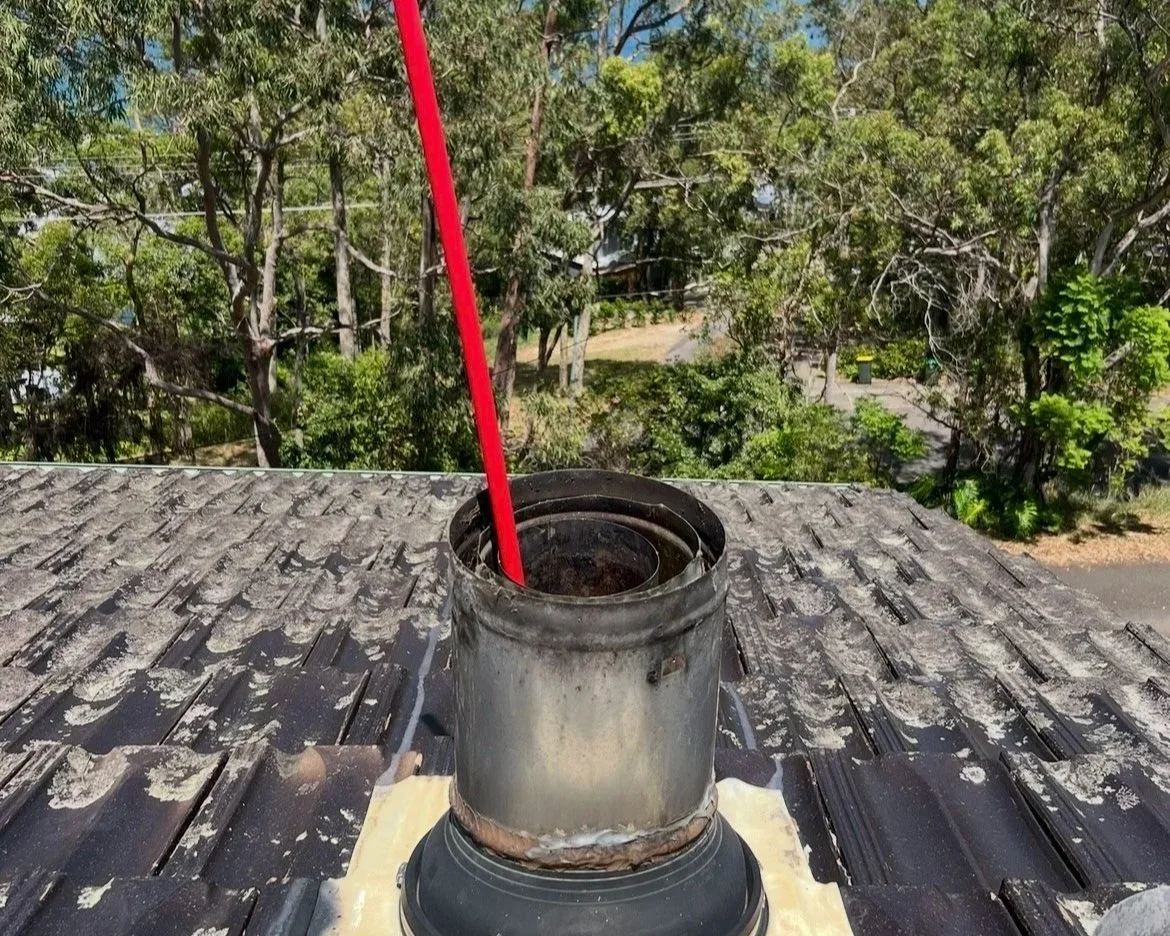 View of a chimney pipe in the process of being cleaned, on a tiled roof with trees in the background.