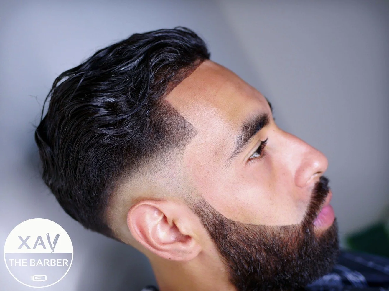 Side profile of a man with styled, slicked-back dark hair and a full beard, lying against a gray background with a logo that reads 'XAV! THE BARBER' in the lower left corner.
