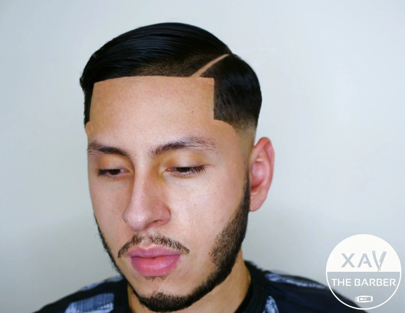 Close-up of a young man with a fresh black hairstyle with a side part and tapered, looking down with closed eyes, in a barbershop setting.