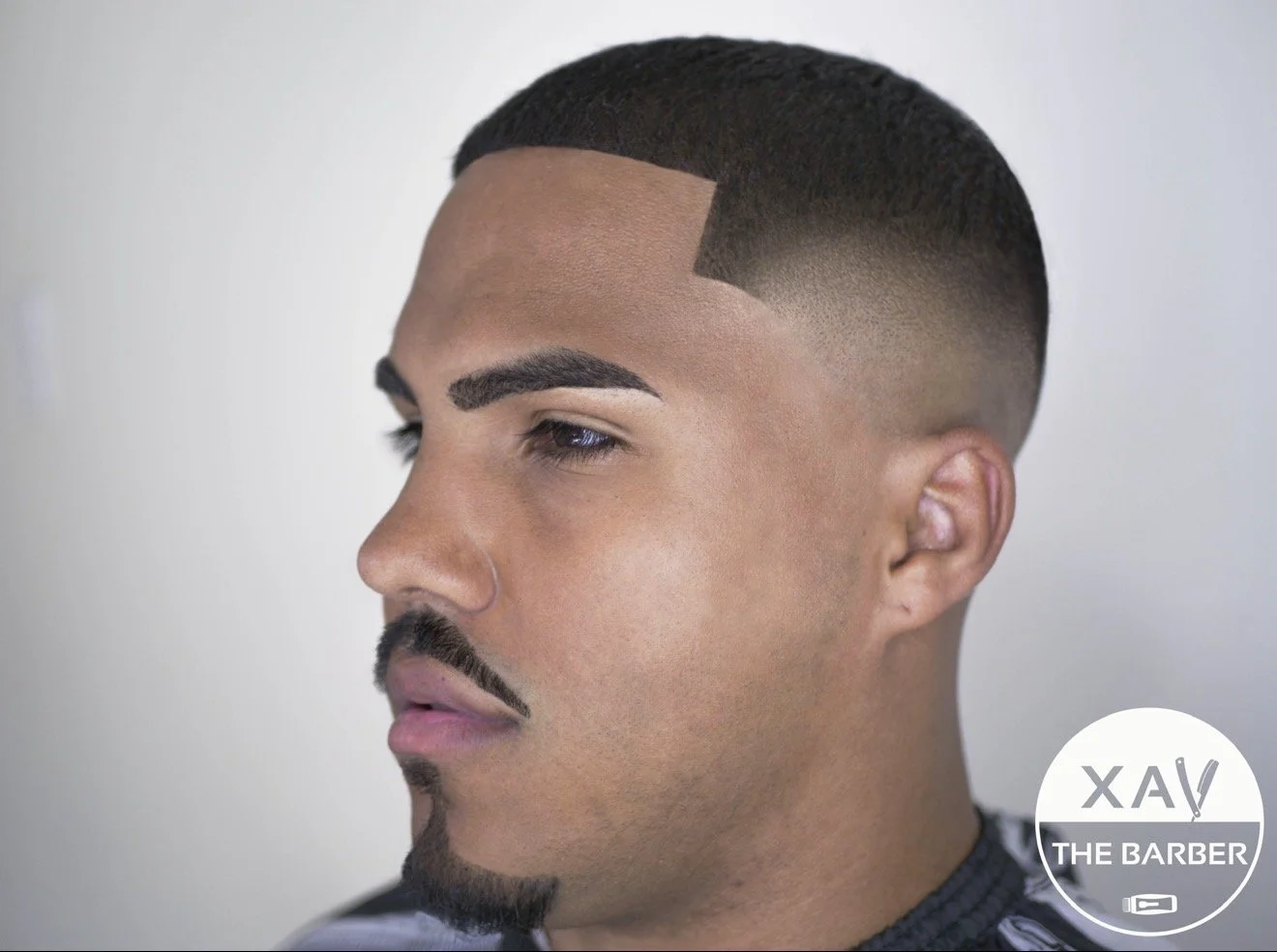 Close-up of a man's face showing a fresh fade haircut, neat beard, and well-groomed eyebrows, with a logo reading 'XAV THE BARBER' in the bottom right corner.