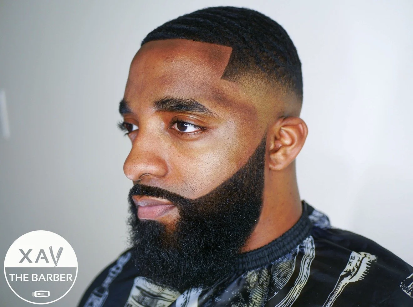 Close-up of a man with a freshly styled haircut and beard, wearing a barber cape, in a barbershop with a plain wall background.