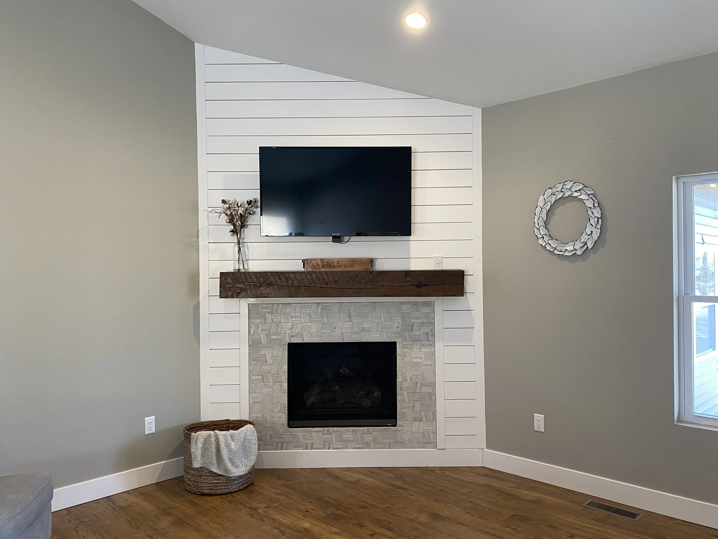 Living room corner with a fireplace, mounted flat-screen TV, decorative shelf, round wall decor, window, woven basket with blanket, and wood flooring.