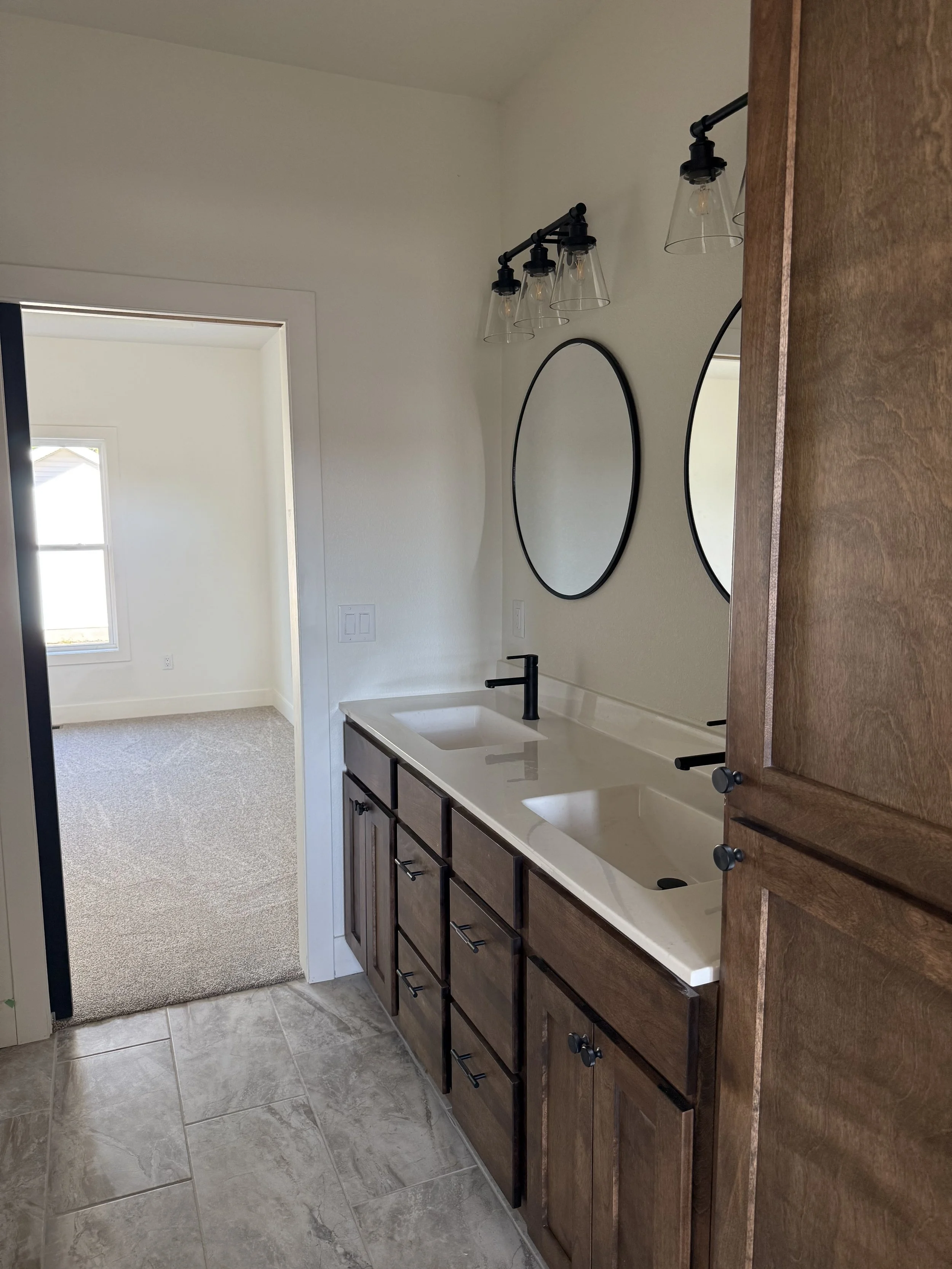 Home bathroom vanity with double sinks, dark wood cabinets, black fixtures, two oval mirrors, and a black light fixture with four exposed bulbs.