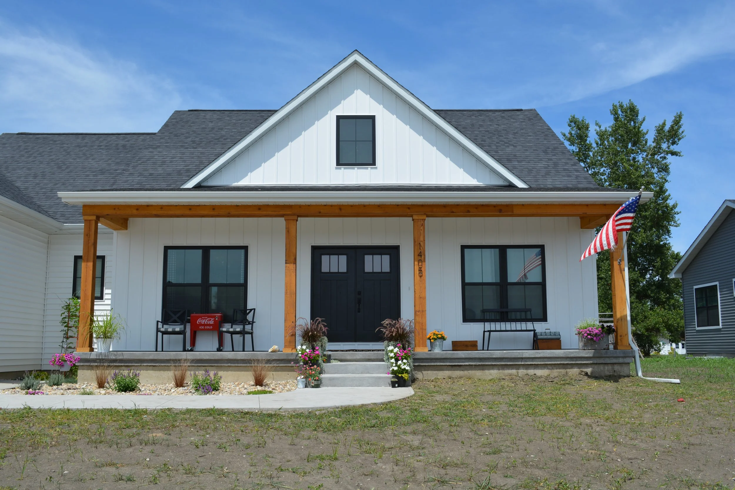 Front view of a modern white house with black front door, two black window frames, a porch with seating, floral decorations, and an American flag, under a blue sky.