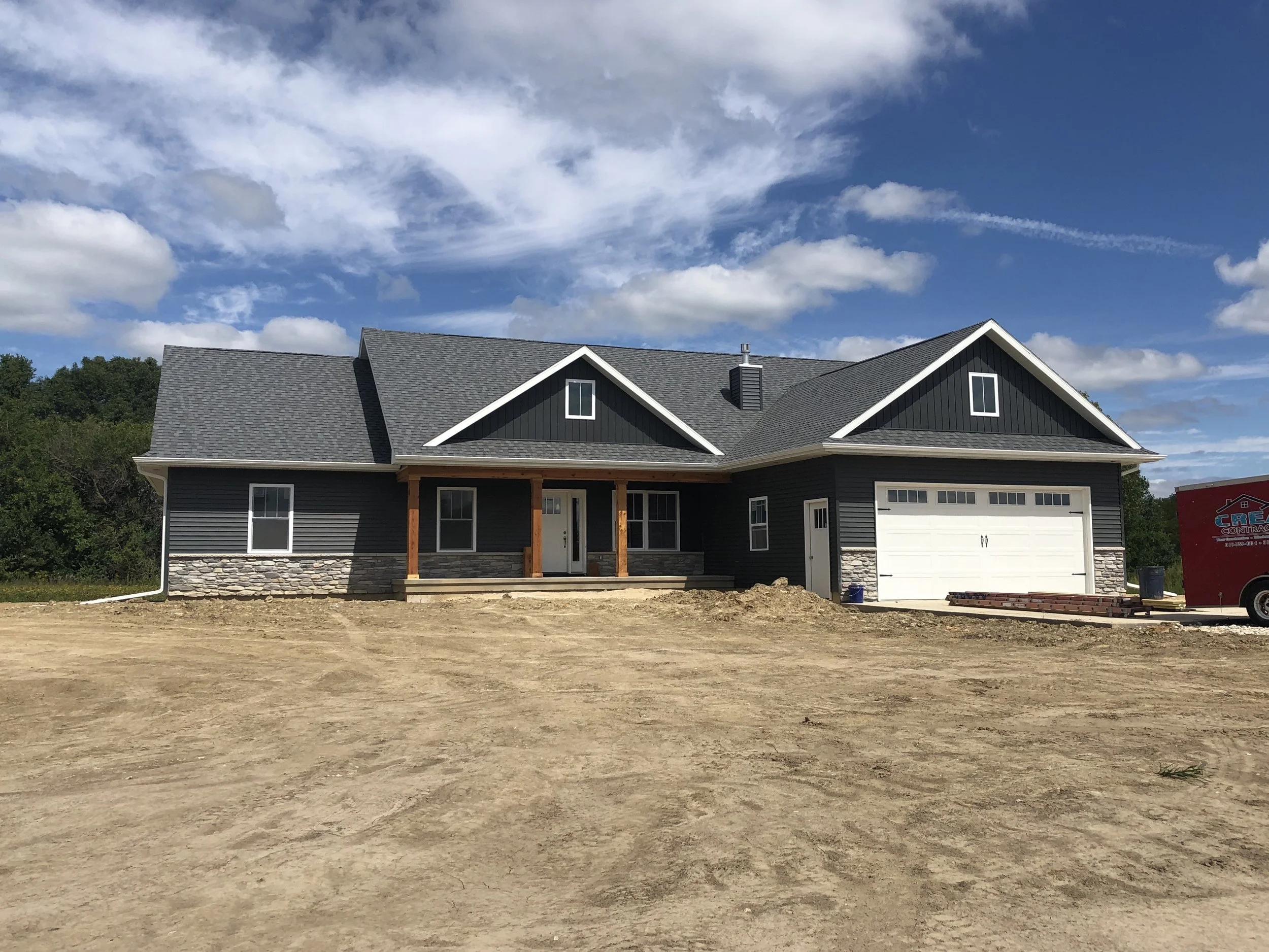 New house under construction, with gray siding, stone accents, and a garage, on a dirt lot under a partly cloudy sky.