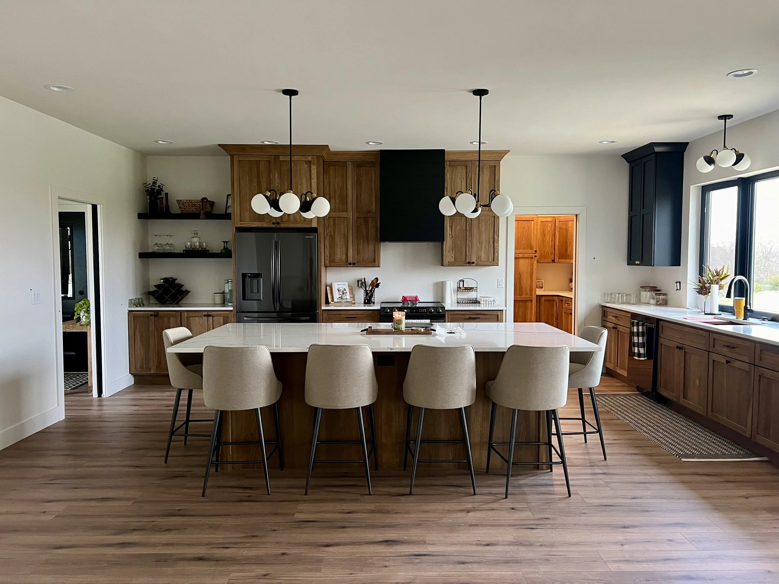 Modern kitchen with wooden cabinets, a large central island with beige chairs, black and white lighting fixtures, stainless steel refrigerator, and hardwood floors.