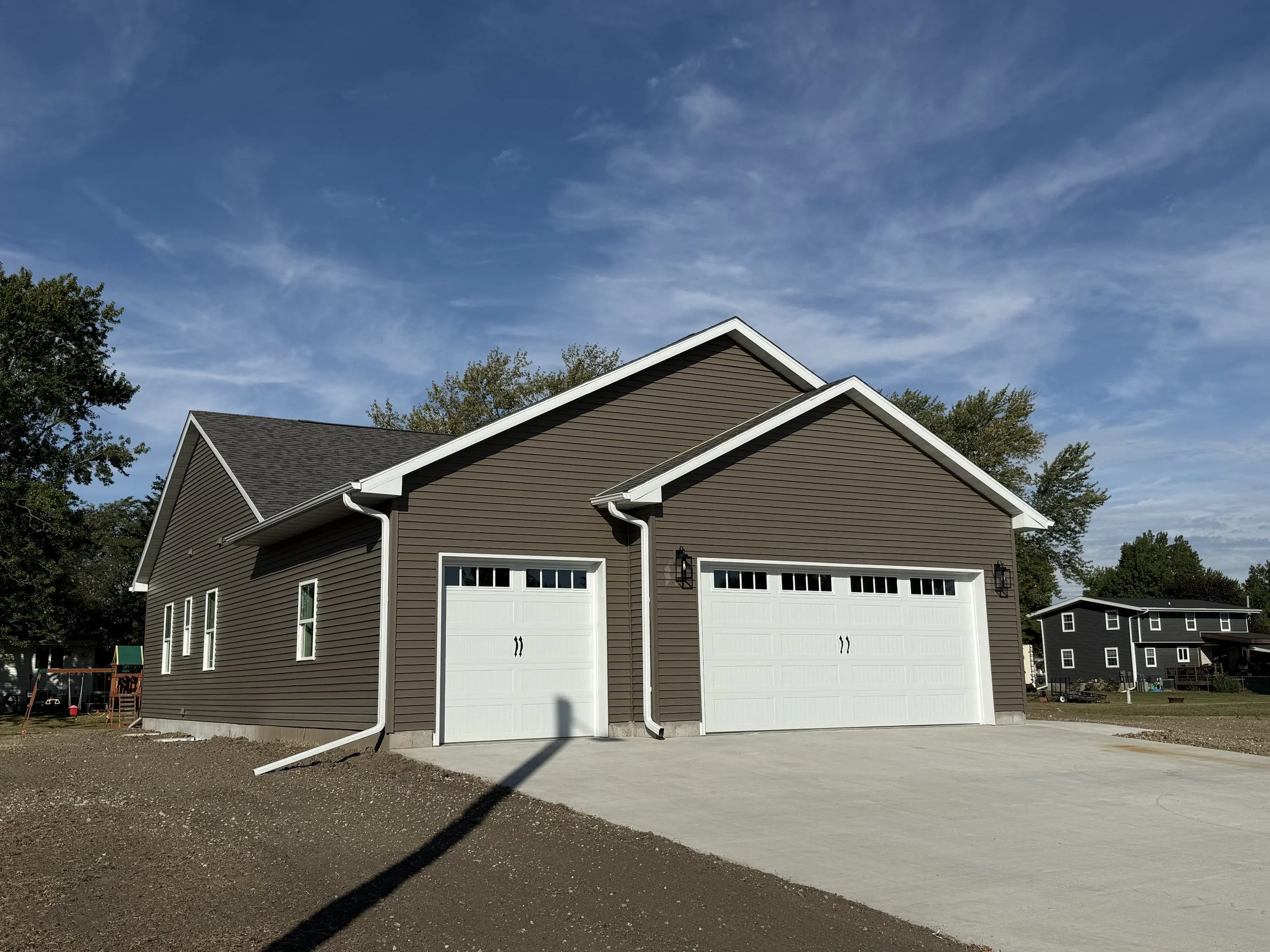 Newly constructed brown house with two white garage doors and a concrete driveway, under a blue sky with some clouds.