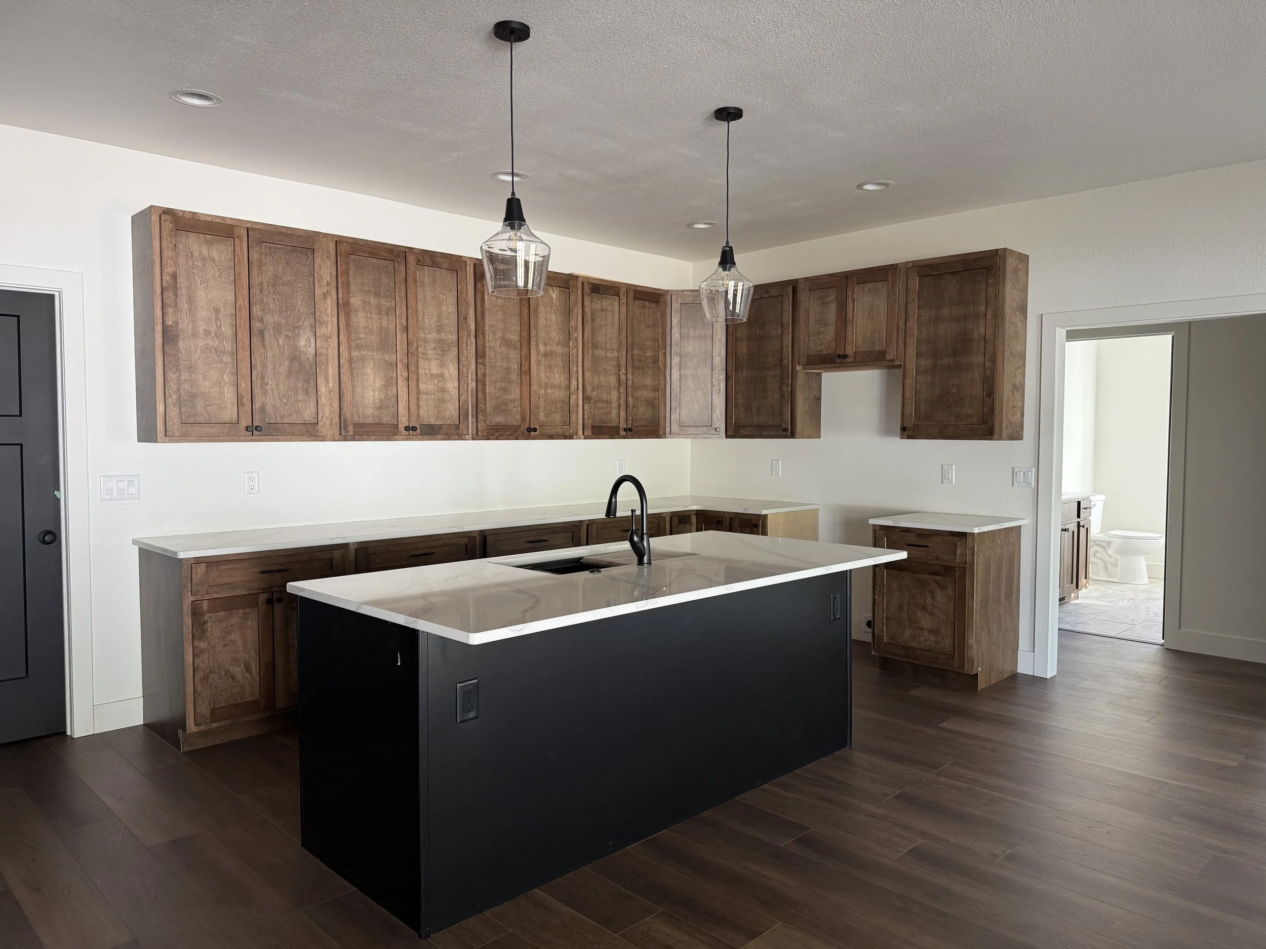 Empty kitchen with wooden cabinets, a black kitchen island with a white marble top, pendant lights, and a doorway leading to a bathroom with a toilet.