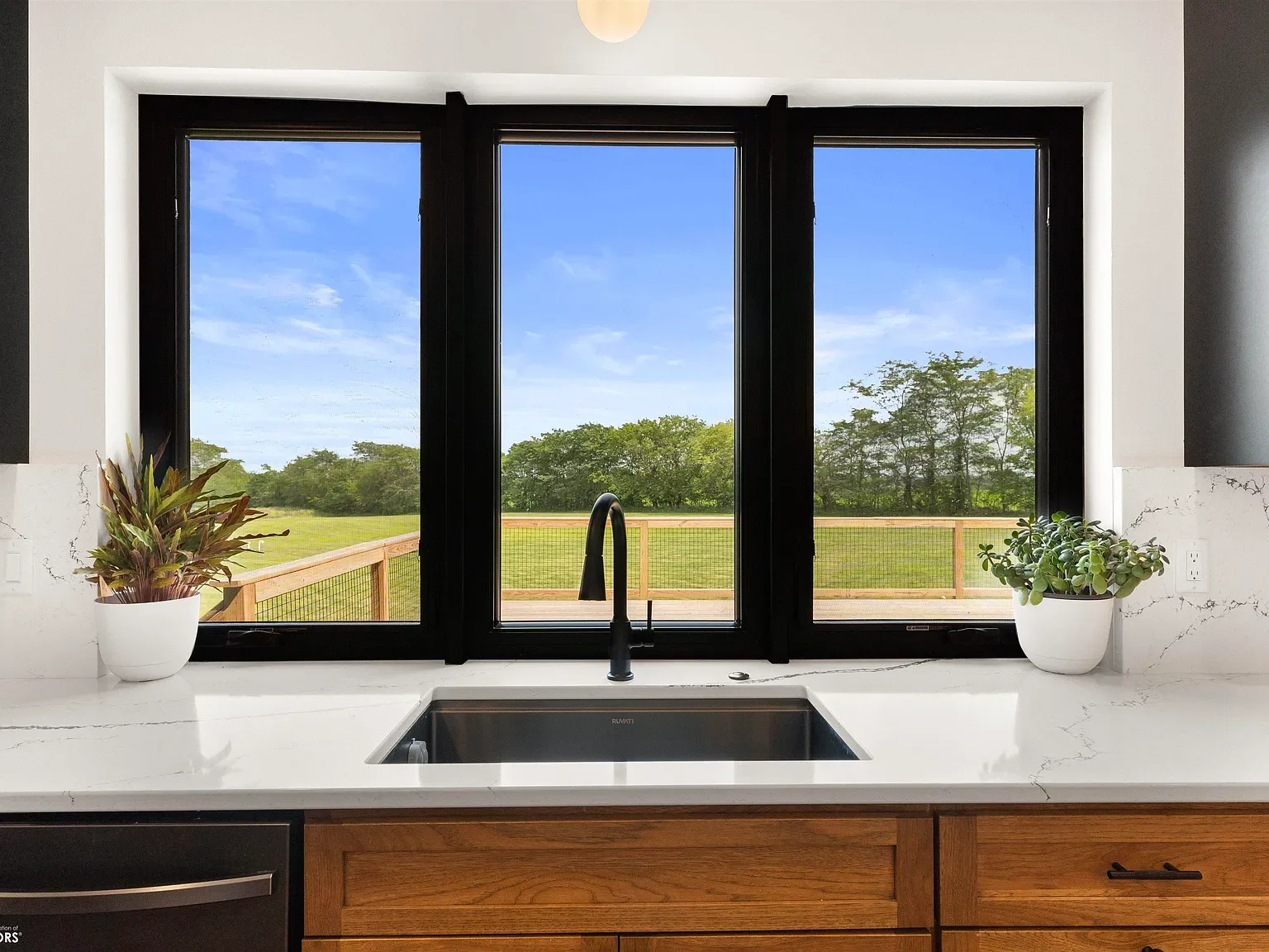 Kitchen sink with white marble countertop, black faucet, and a view of a grassy field and trees through a large window with black framing, flanked by potted plants on both sides.