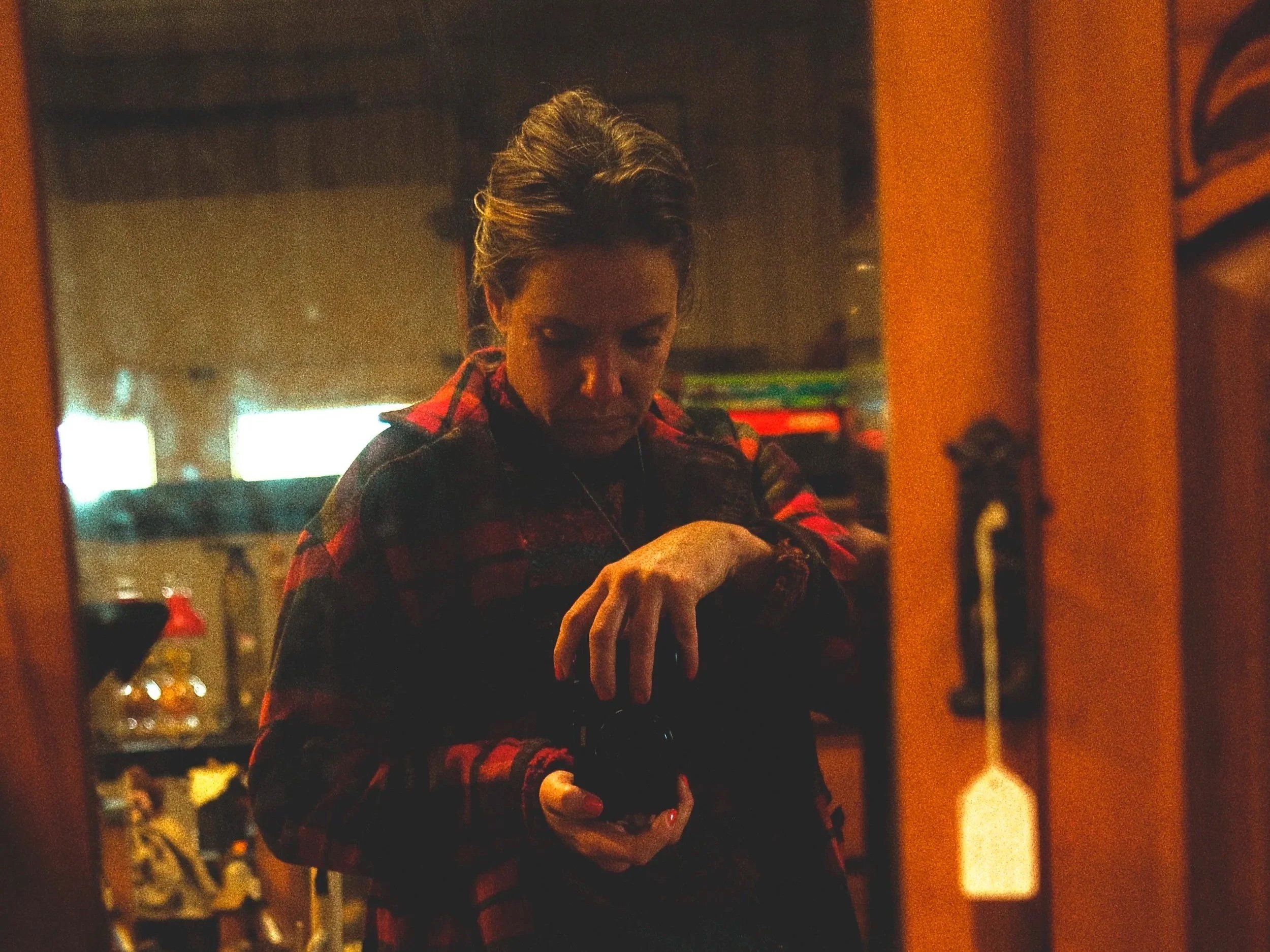 A woman taking a selfie in front of a mirror, holding a camera or phone, wearing a red and black plaid shirt, in a dimly lit room with warm lighting.
