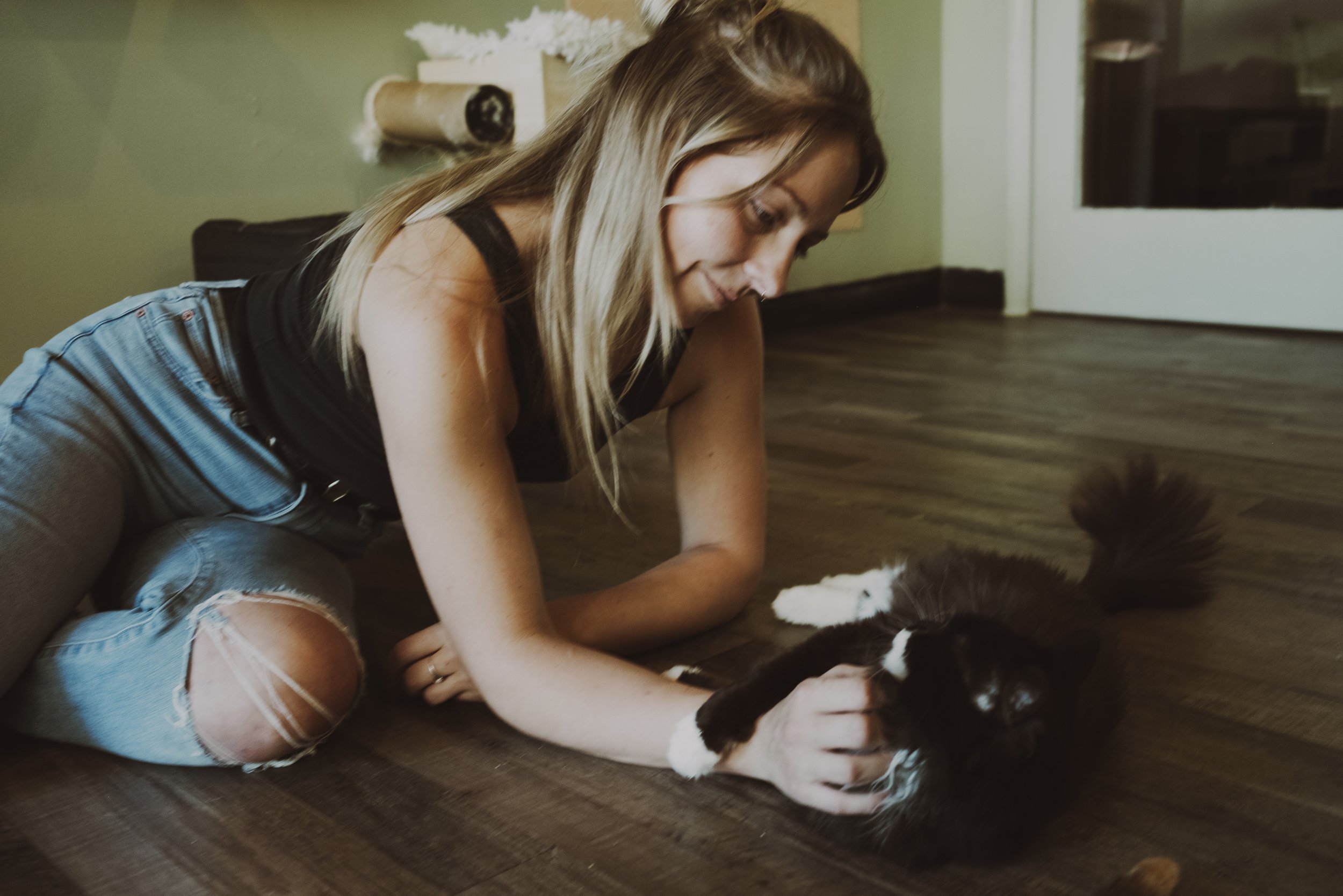 A girl with long blonde hair, wearing a black tank top and ripped jeans, playing with a black and white cat on a wooden floor inside a home.