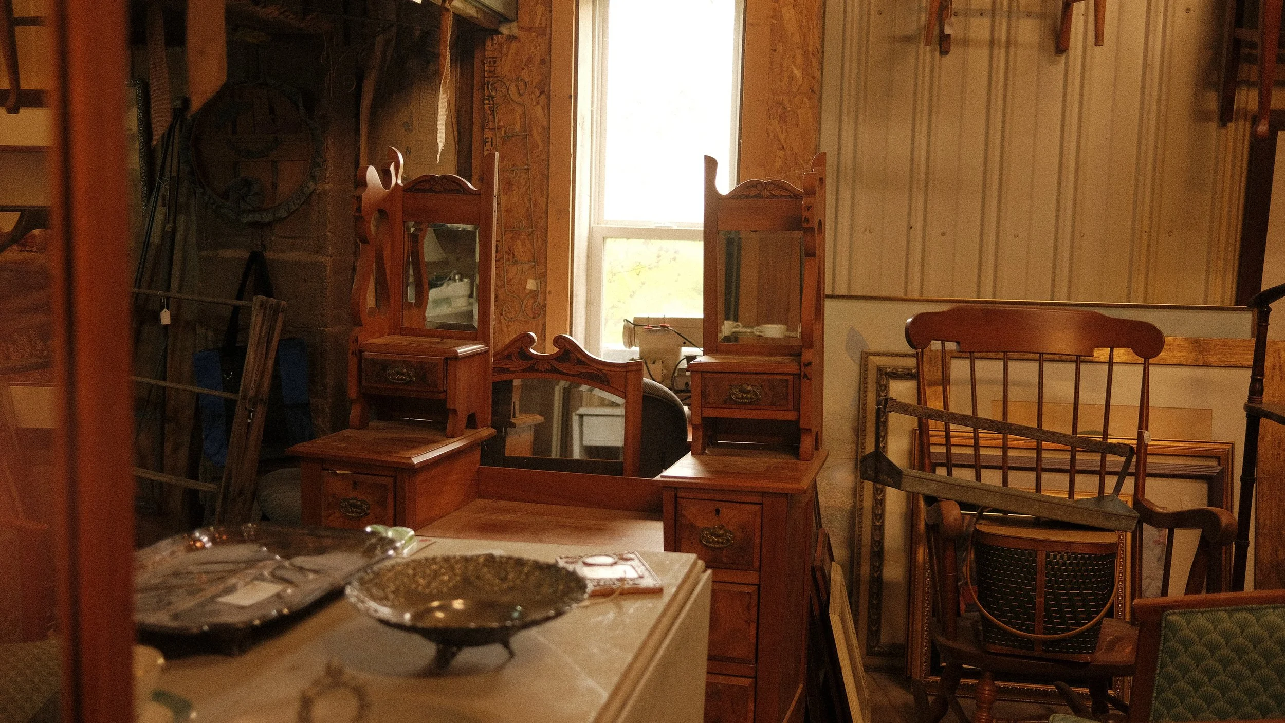 A collection of antique wooden furniture, including dressers, chairs, and a vanity mirror, arranged in a cluttered room with a window in the background.
