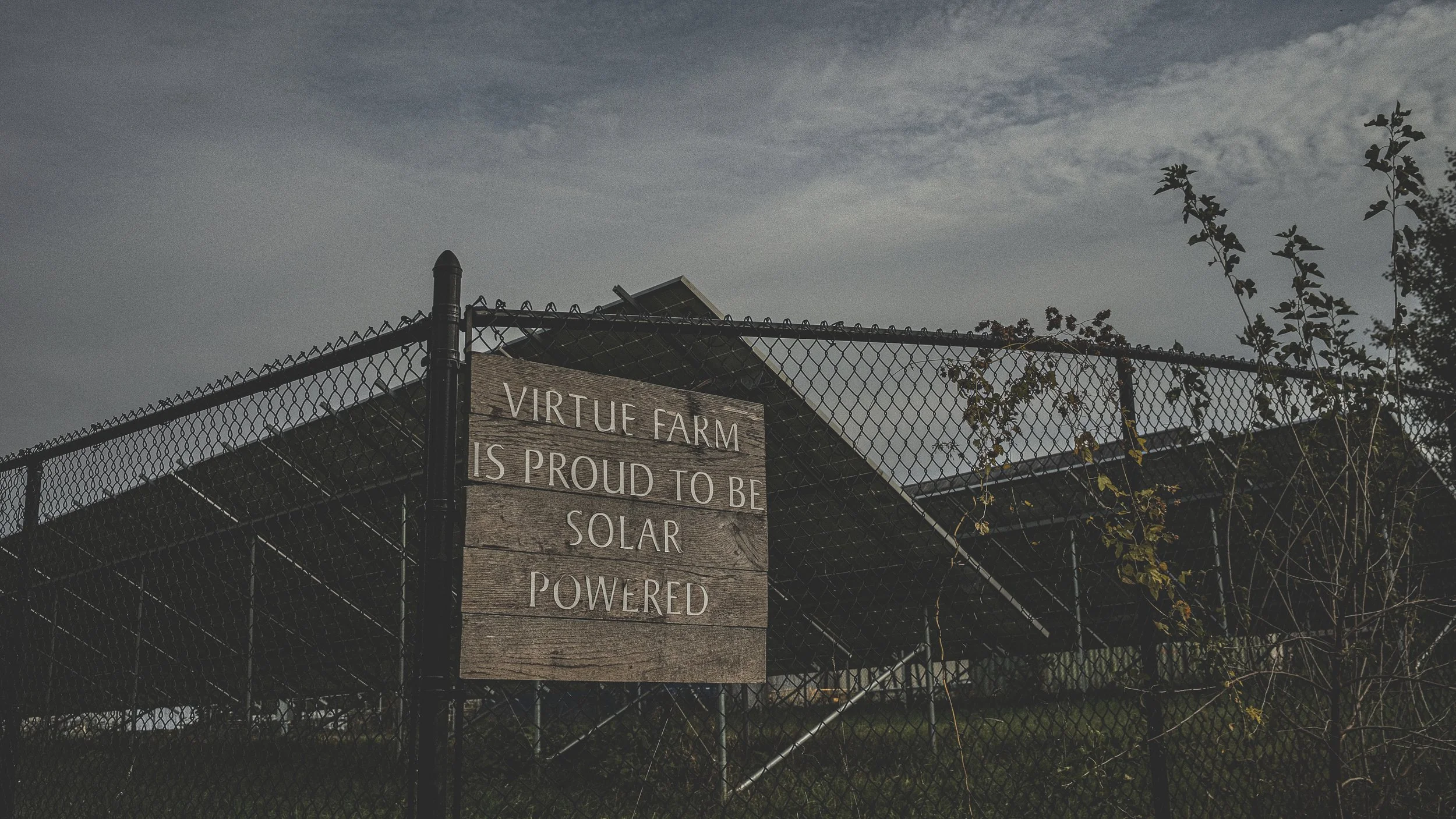 Wooden sign on fence reads 'Virtue Farm is proud to be solar powered.' Behind the fence, solar panels and farm structures are visible under a cloudy sky, with some trees and bushes nearby.