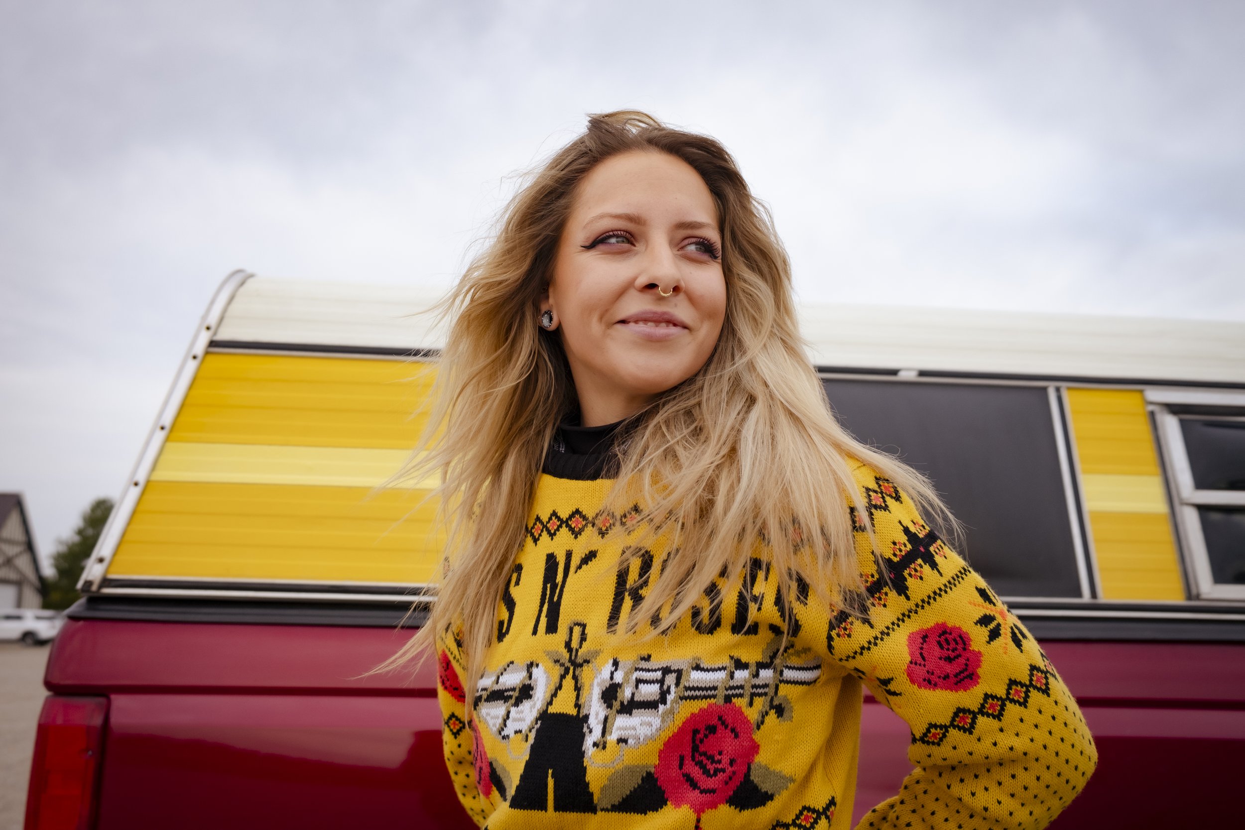A woman with blonde hair, piercings, and earrings, wearing a yellow sweater with red roses and black and white designs, stands in front of a maroon vehicle with a yellow and black camper attached. She is looking to the side with a slight smile.