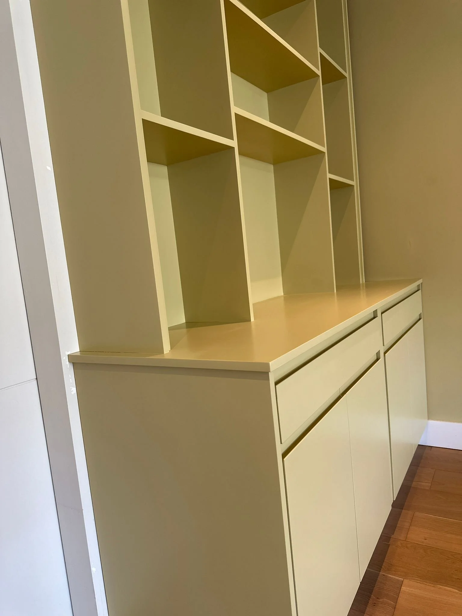 A cream-colored built-in cabinet with open shelving on top and inlaid drawers and cabinets at the bottom, against a light-colored wall and next to a wooden floor.