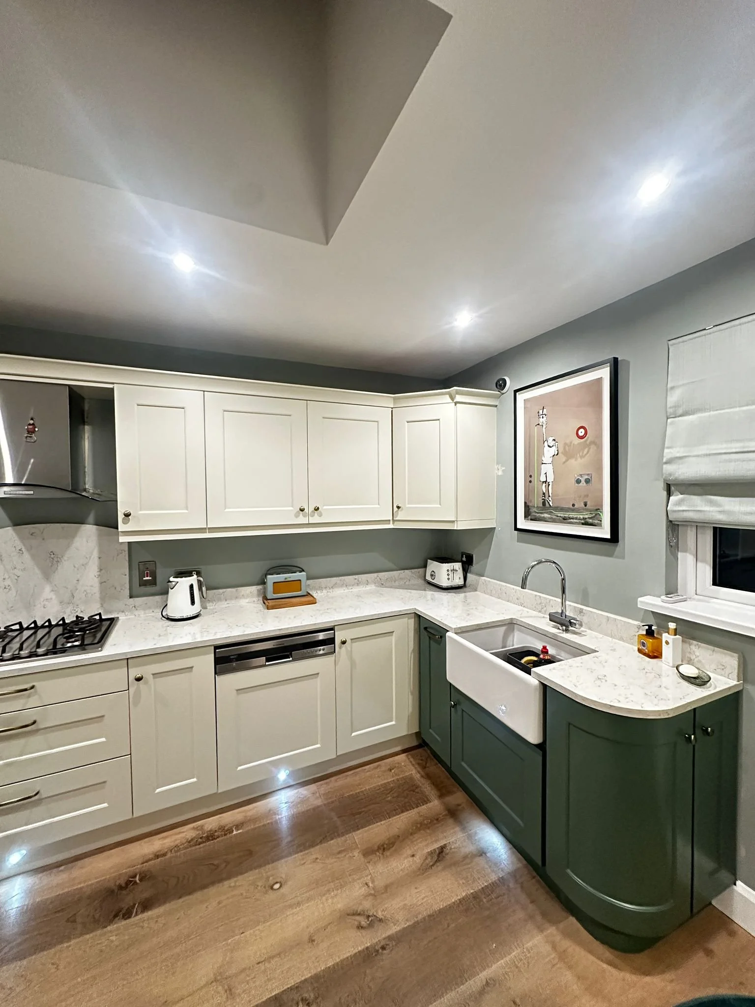 Modern kitchen with white upper cabinets and green lower cabinets, marble countertops, a white farmhouse sink, wooden flooring, and a framed artwork on the wall.