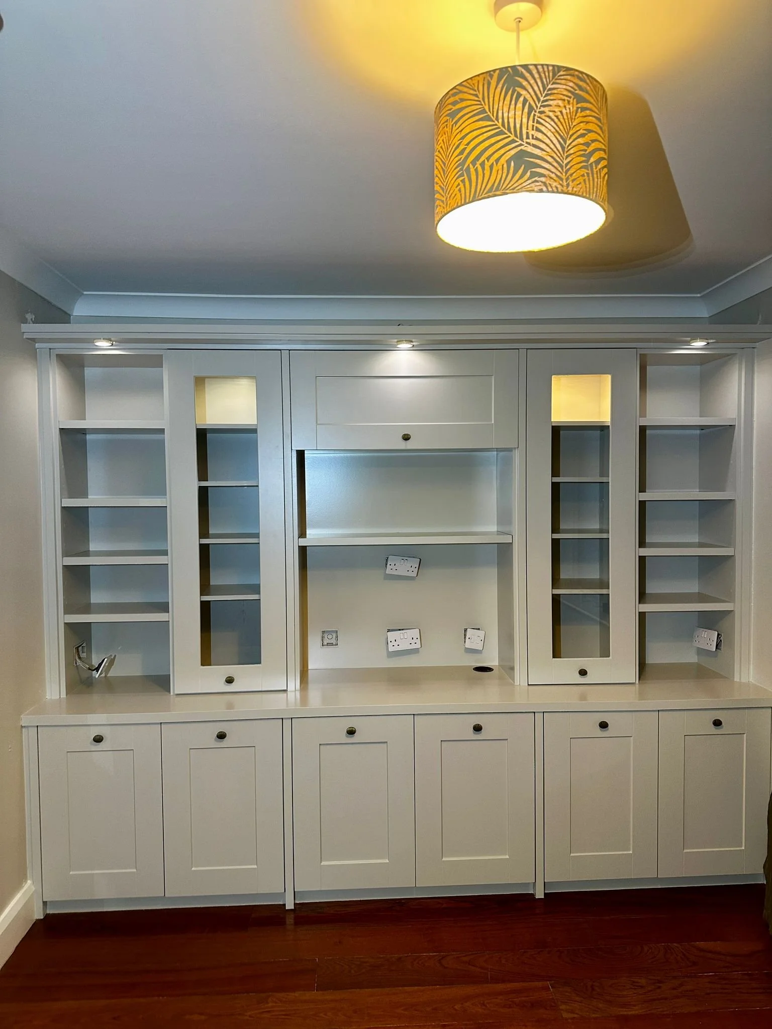 Empty built-in white cabinetry with open shelves and closed cabinets below, electrical outlets, and a ceiling light with a yellow patterned lampshade.